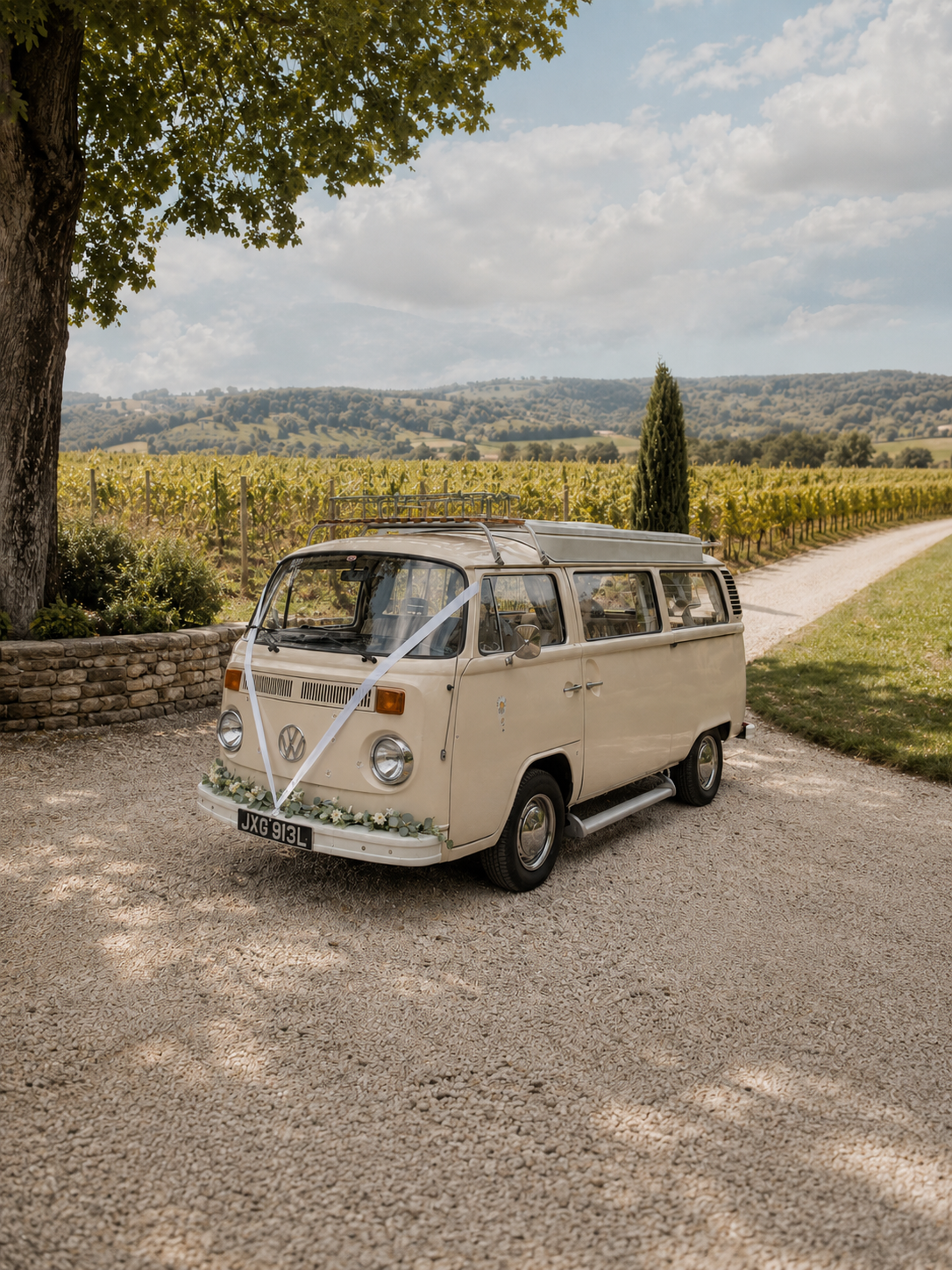 A vintage Volkswagen van decorated with flowers, parked on a gravel pathway beneath a large tree, with a vineyard and rolling hills in the background. Vintage wedding car for a real wedding in West Sussex