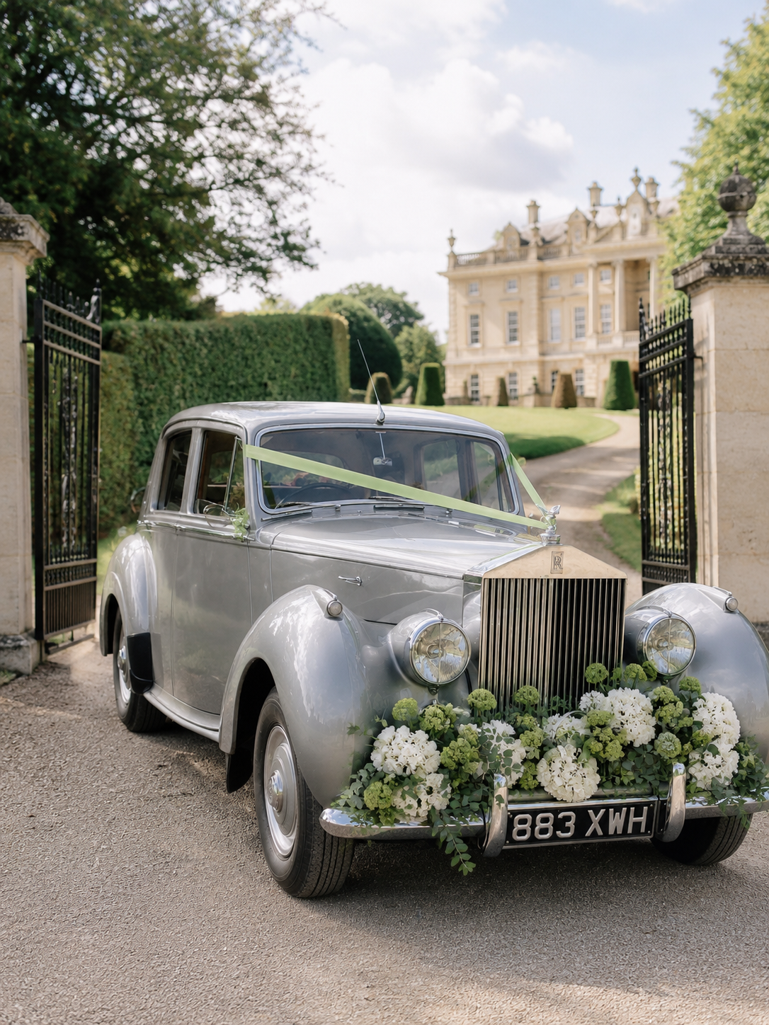 A vintage silver luxury car decorated with white and green flowers, parked at the entrance of a grand estate with a large mansion in the background.