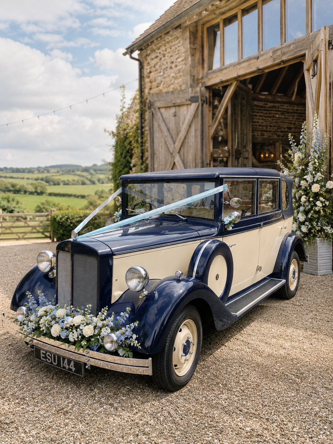 A classic vintage car decorated with white roses and blue ribbons, parked outside a rustic barn with open doors, against a backdrop of green fields and a partly cloudy sky.