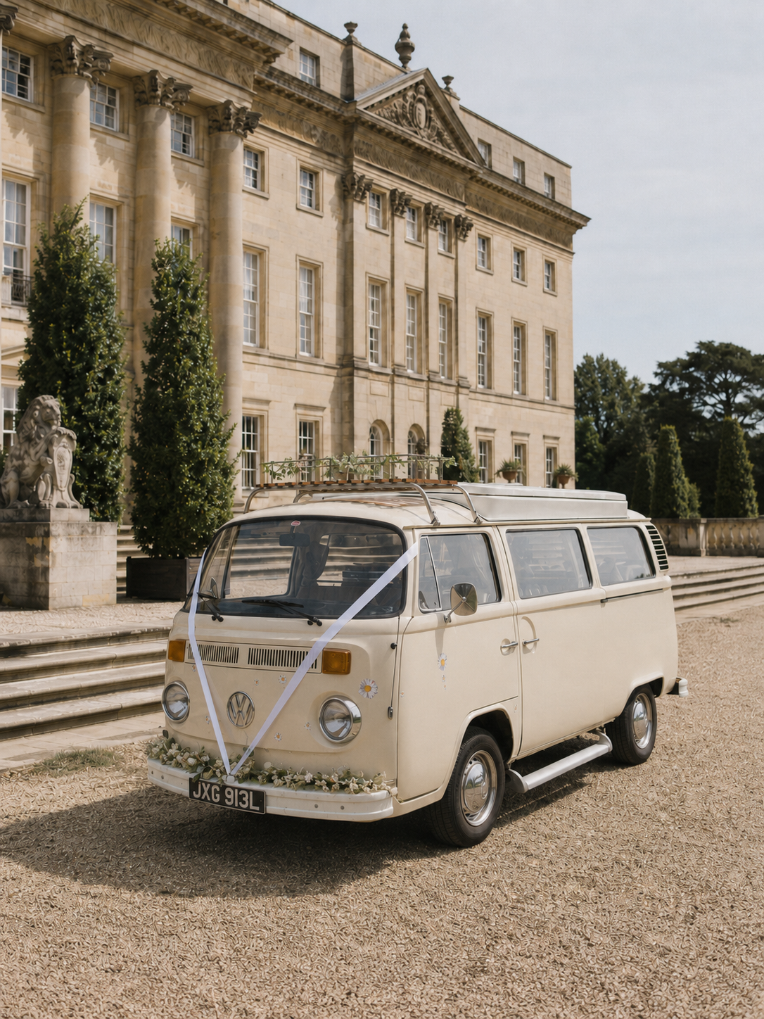 A vintage beige Volkswagen camper van with wedding decorations parked in front of an elegant stone mansion with steps and columns, surrounded by greenery.
