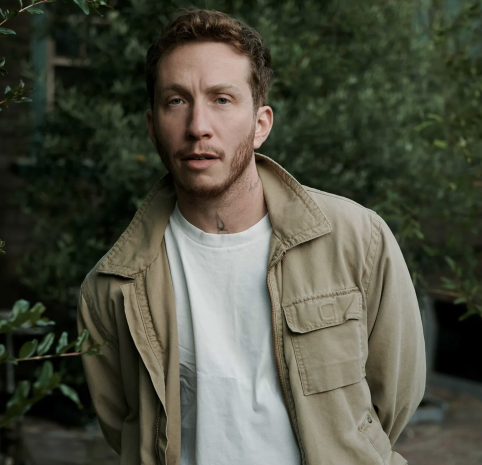A man with curly brown hair and a beard, wearing a beige jacket over a white t-shirt, standing outdoors among green bushes.