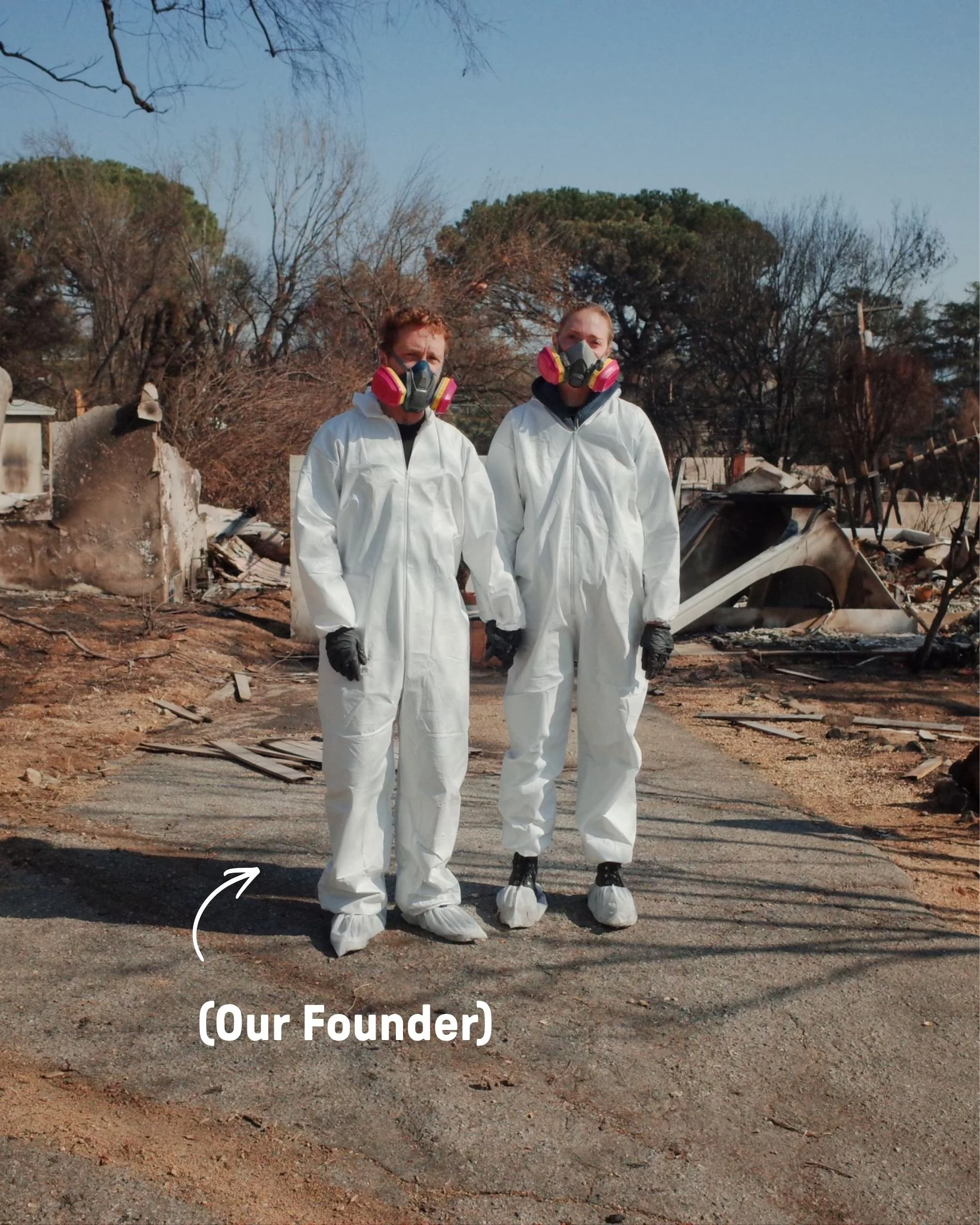 Two individuals wearing white protective suits and respirator masks standing in a damaged area with debris and burnt trees, likely after a disaster, with a background of blue sky and trees.
