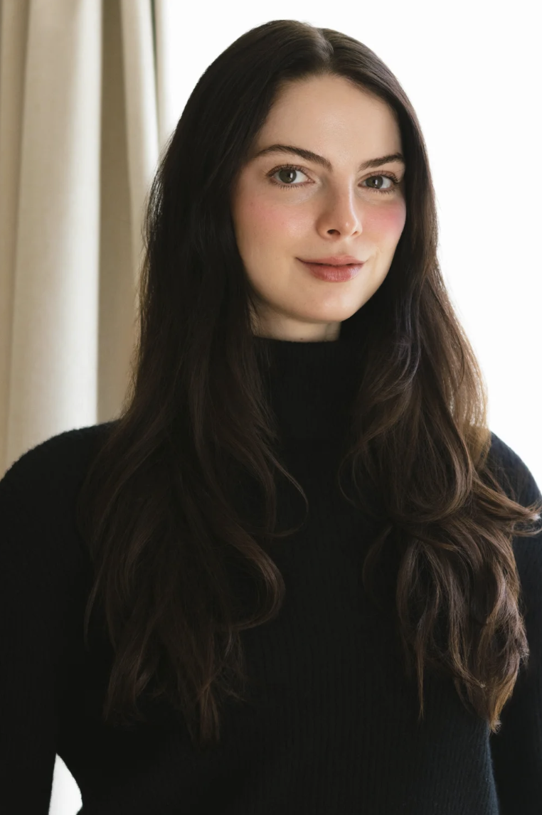 A young woman with long brown hair, wearing a black turtleneck sweater, standing indoors near a light-colored curtain with natural light coming from the side.