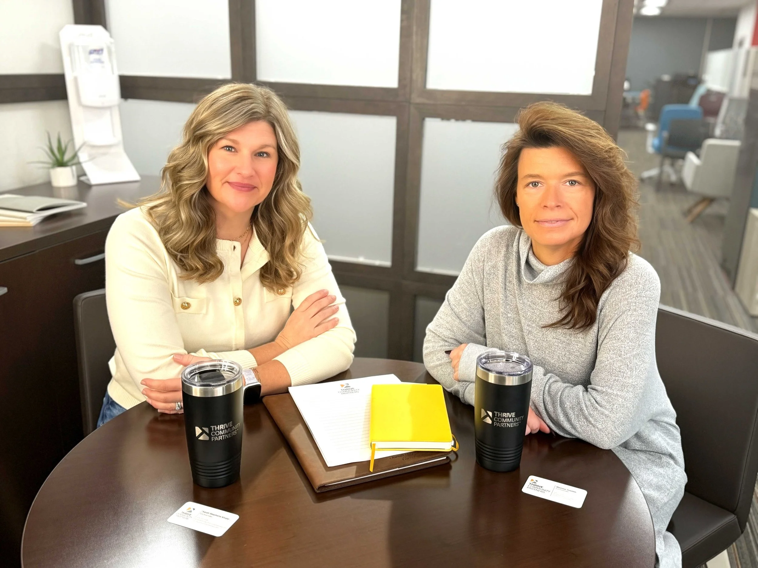 Two women sitting at a round table with notebooks and travel mugs, smiling at the camera in an office setting.