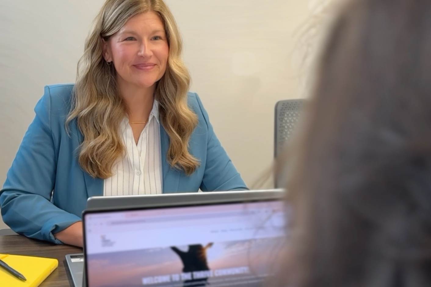 A woman with long wavy blonde hair wearing a blue blazer and white blouse sitting at a desk, smiling during a conversation with an unseen person in an office setting.