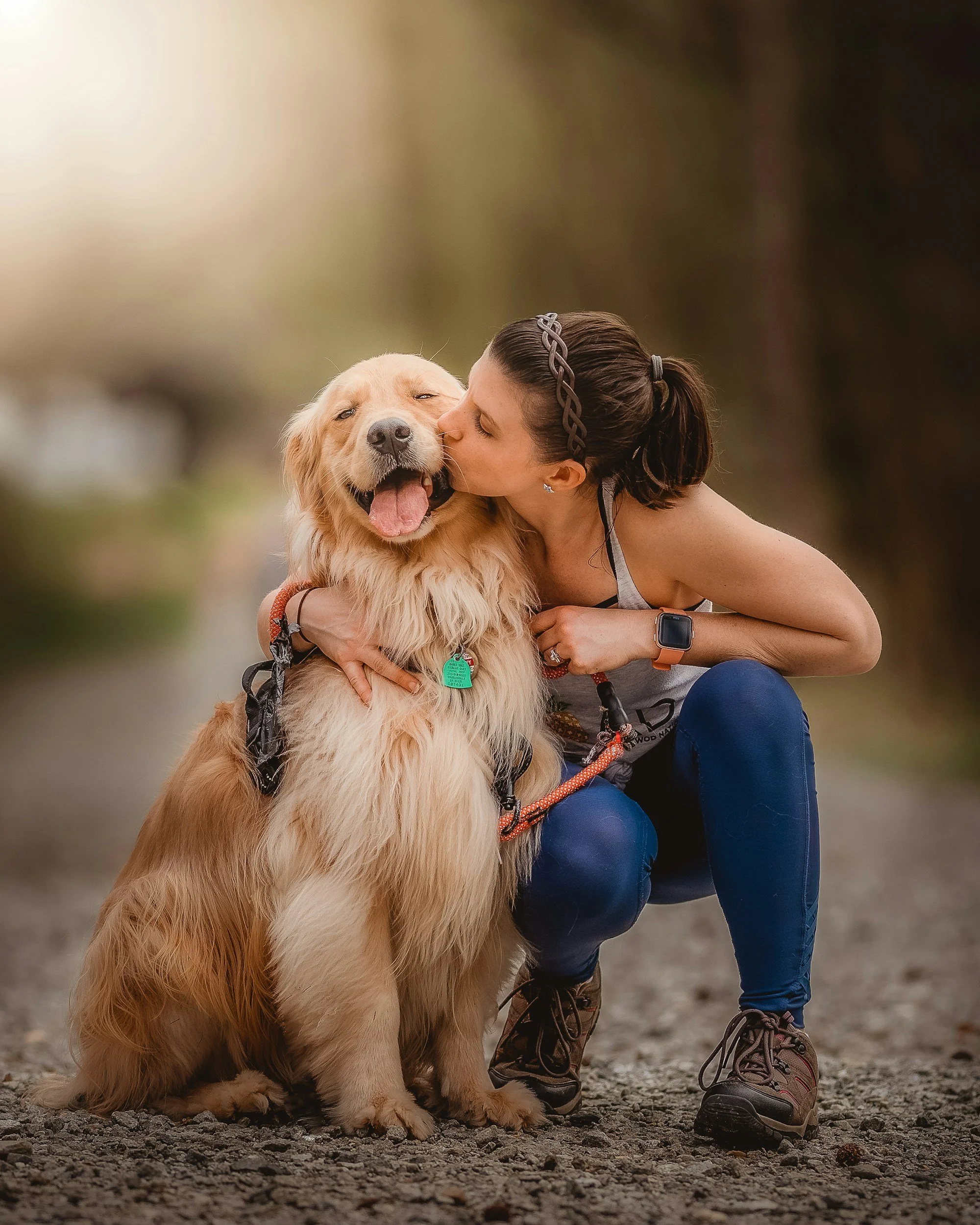 A woman with brown hair tied in a ponytail, wearing a grey tank top, blue jeans, brown hiking boots, and an Apple Watch, is hugging and kissing a large, golden retriever dog with a collar and a green tag, on a gravel surface outdoors.