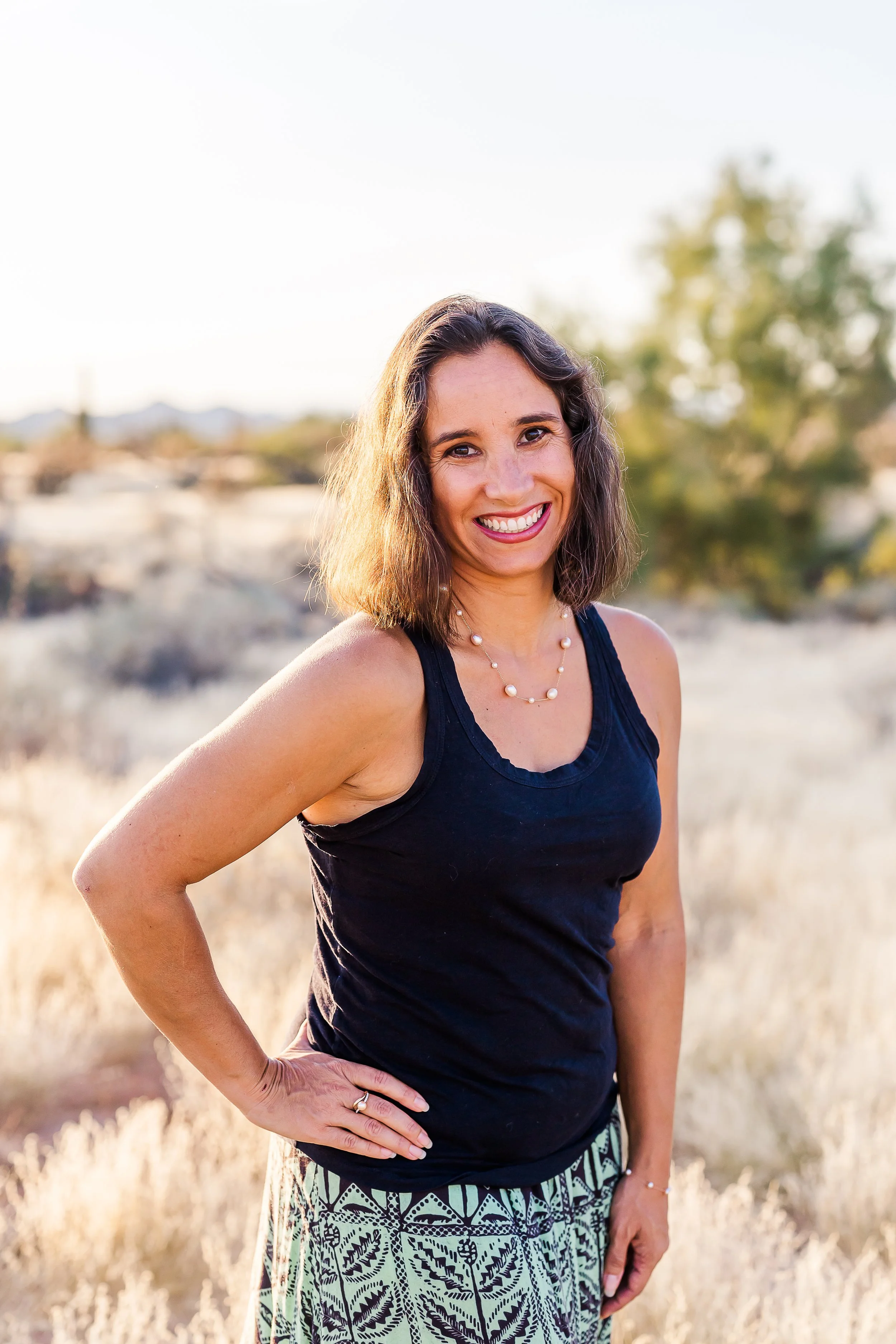 A woman standing outdoors in a field of dry grass, smiling at the camera with her hand on her hip, wearing a black sleeveless top and patterned skirt, with trees and mountains in the background during sunset.