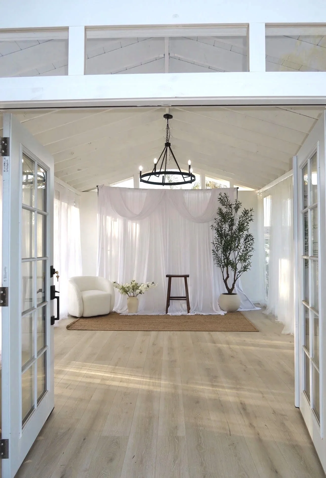 Interior porch with open double doors, white curtains, a white chair, a small table with flowers, a large potted plant, a woven rug, and a black chandelier