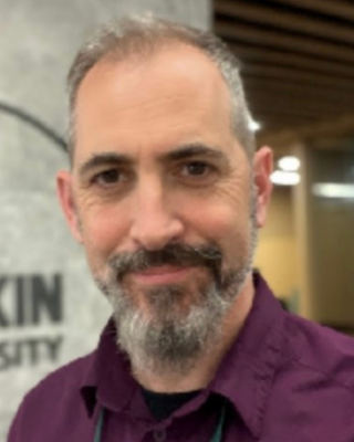 Close-up of a man with gray hair and beard smiling indoors, wearing a purple shirt.