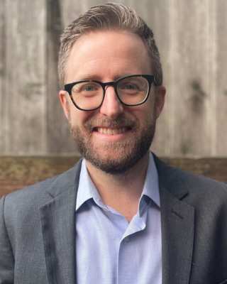 Portrait of a man with glasses, a beard, and gray hair, smiling, wearing a gray blazer and a light blue shirt, standing outdoors in front of a wooden fence.