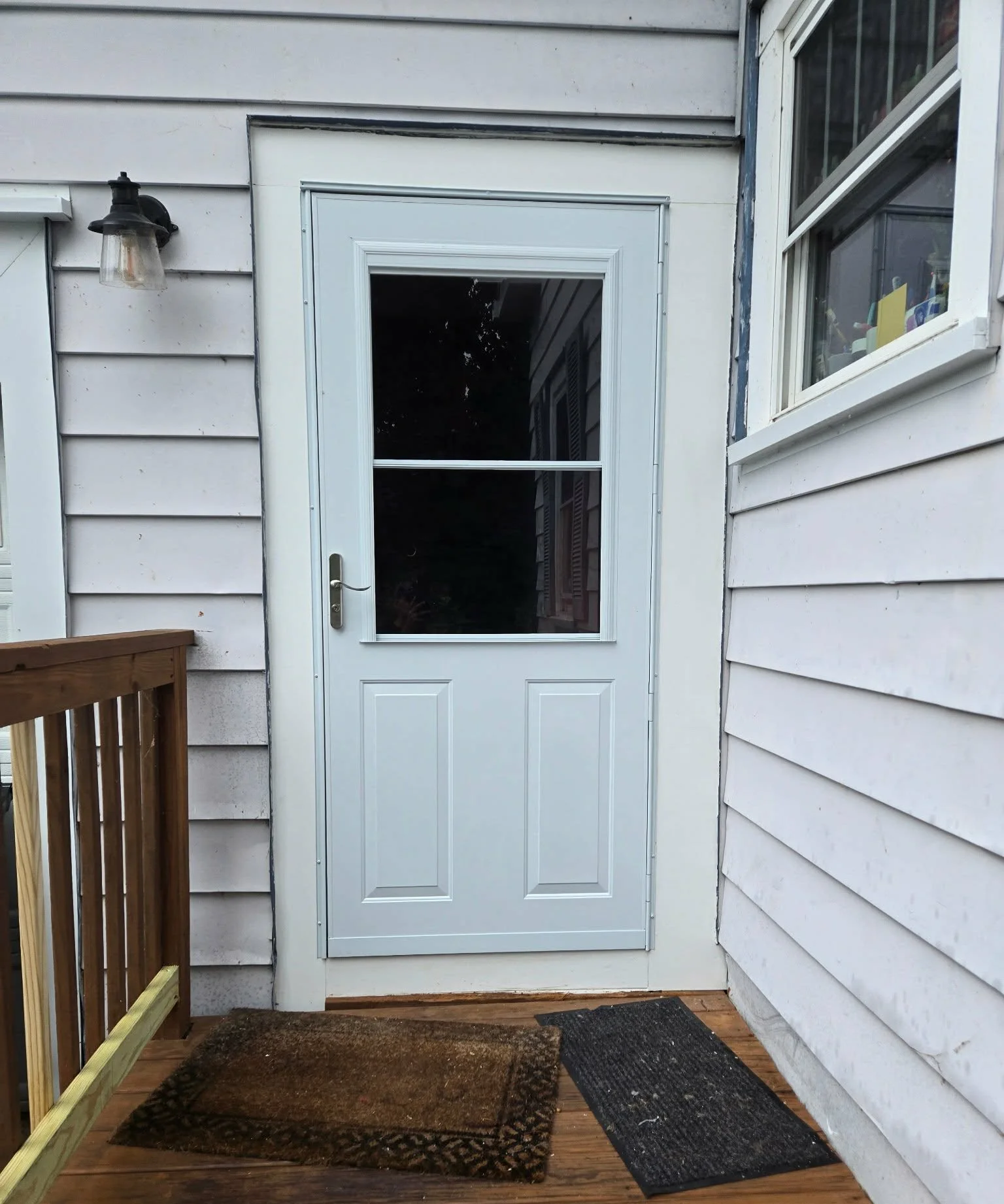 Back door with white frame and glass window, two doormats on wooden porch, white weathered siding, outdoor wall lantern, partially visible wooden railing on left, window with items inside on right.