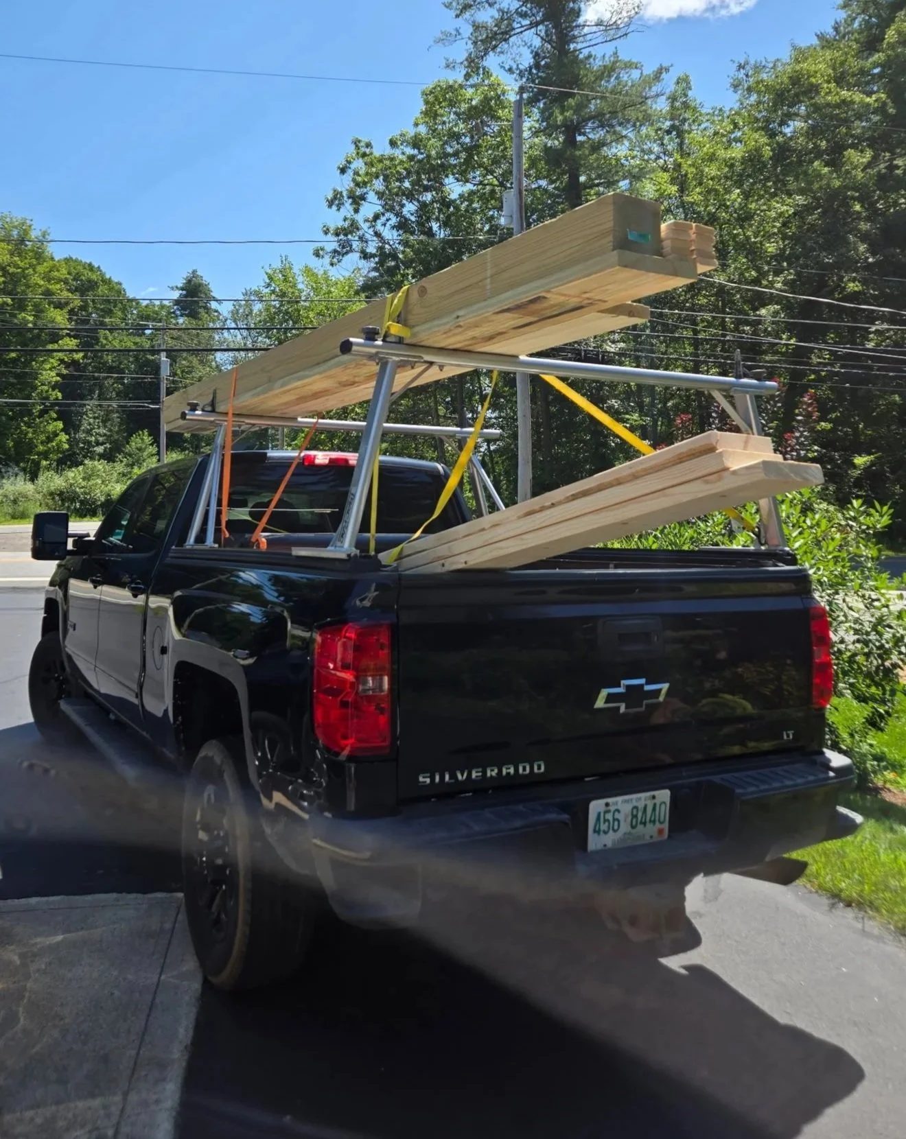 Black Chevrolet Silverado pickup truck parked by the roadside with long wooden planks and beams secured on the truck bed, surrounded by green trees and a blue sky.