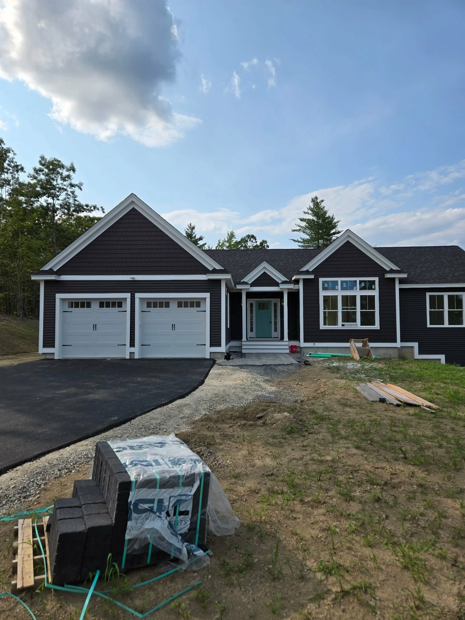 A newly built house with black siding and white trim under a blue sky with some clouds. The house has three garage doors and a front entrance door. Construction materials and tools are on the ground in front of the house, indicating ongoing construction or landscaping work.