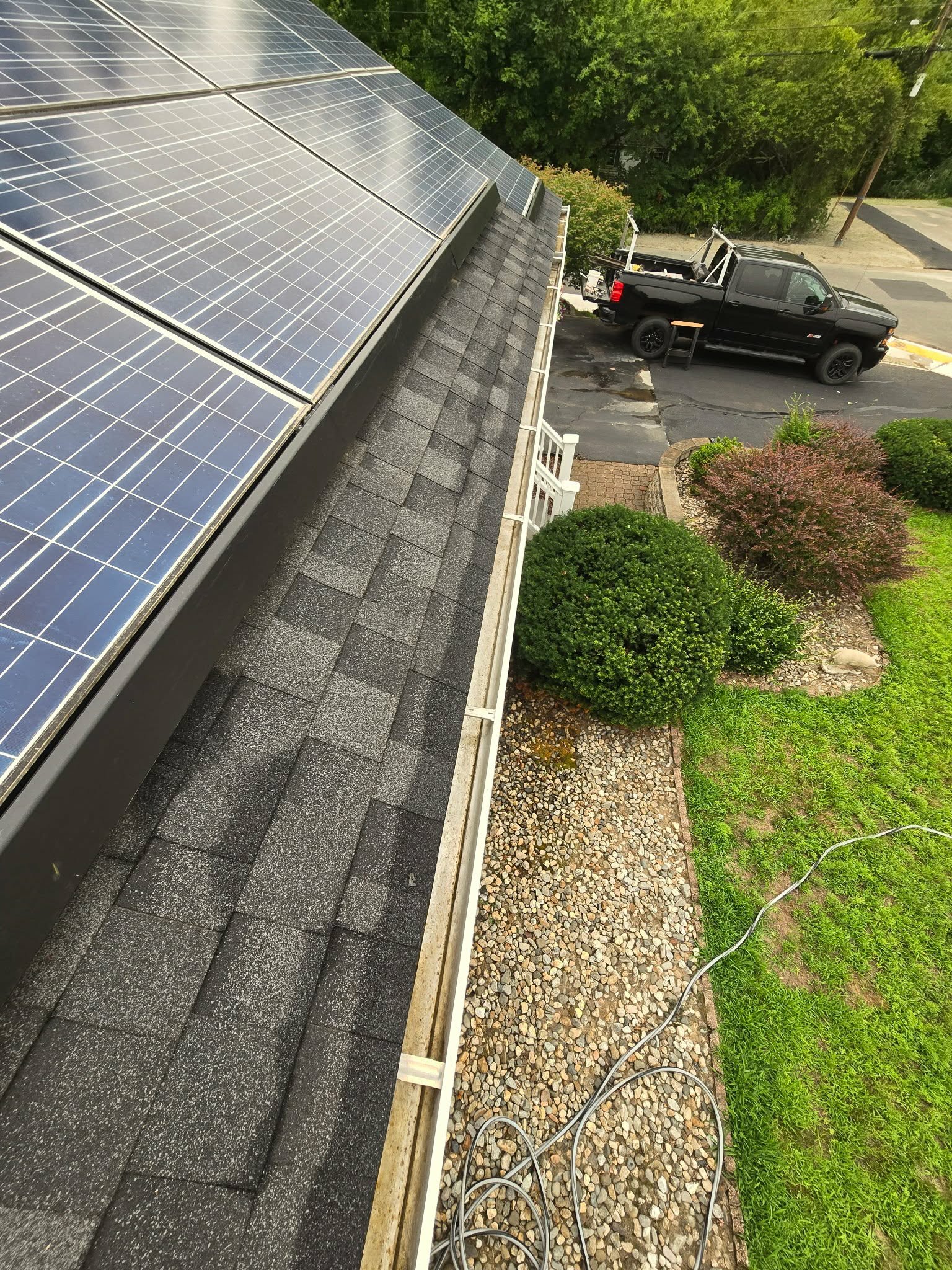 View of solar panels installed on a house roof, with a driveway, a black pickup truck, and trees in the background.