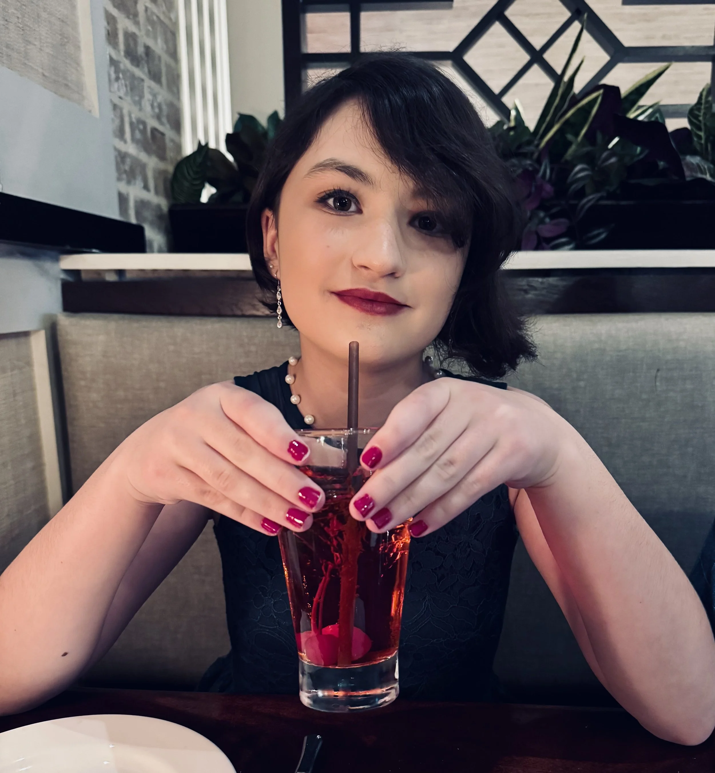 A woman with short dark hair and earrings sitting at a table in a restaurant, holding a glass of red beverage with a straw. She is wearing a black lace dress and has red nail polish. There are plants and a brick wall in the background.