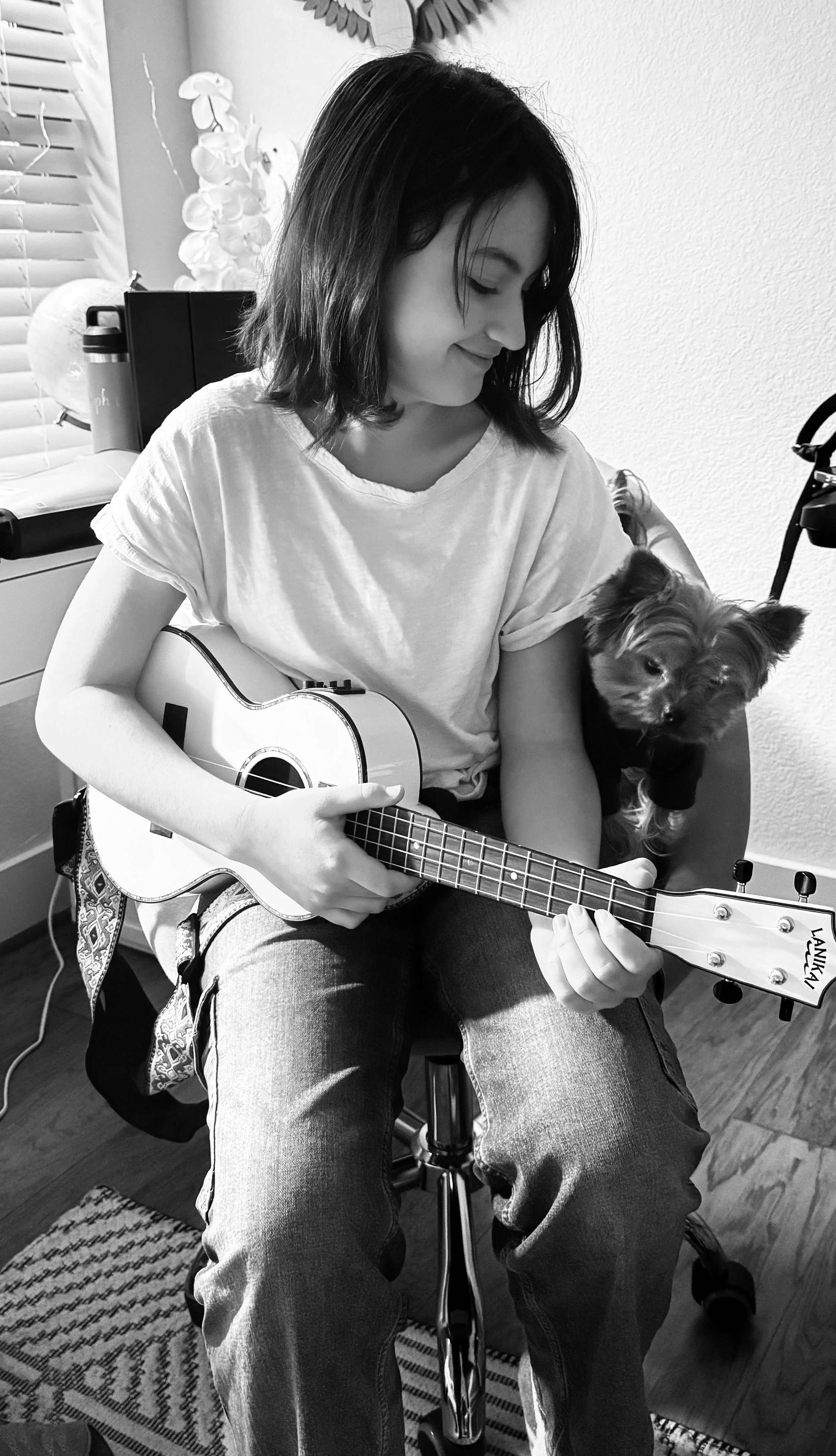 A woman playing a guitar while sitting on a chair with a dog sitting next to her, in a room with blinds and decorative items.