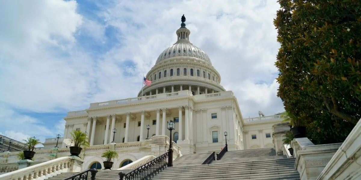 The United States Capitol building in Washington, D.C., seen from the steps leading up to it, with a partly cloudy sky in the background and a large tree on the right side.