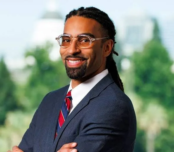 Portrait of a man in a business suit with glasses, smiling, with a blurred outdoor background.