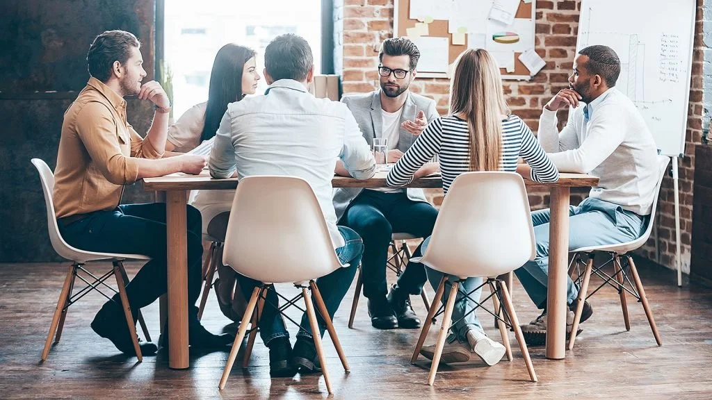 Seven people sitting around a wooden table in a meeting room with exposed brick wall, whiteboard, and bulletin board.