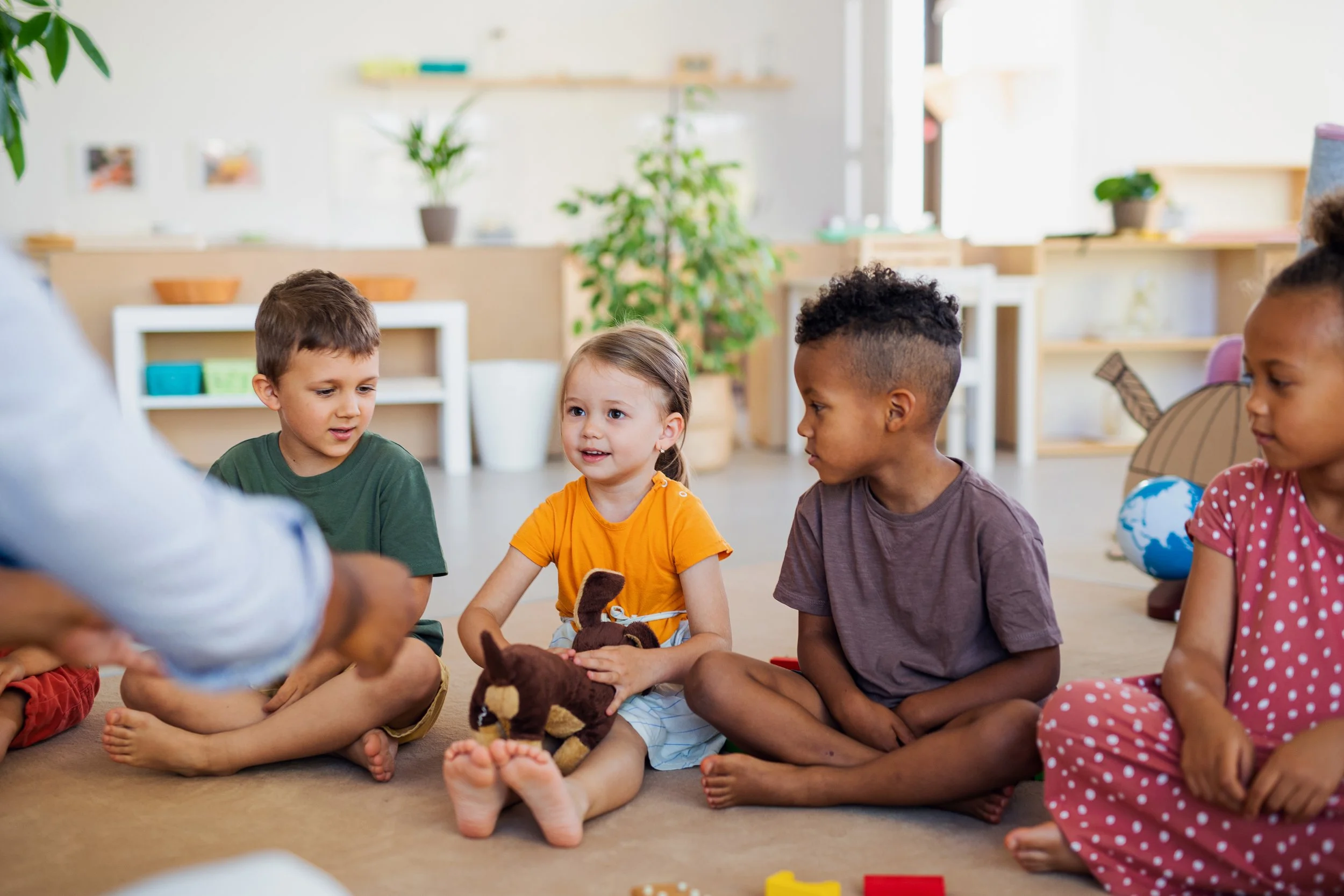 Children sitting cross-legged on the floor engaging in a group activity, with a teacher or adult assisting. The room is bright with decorative items and plants in the background.