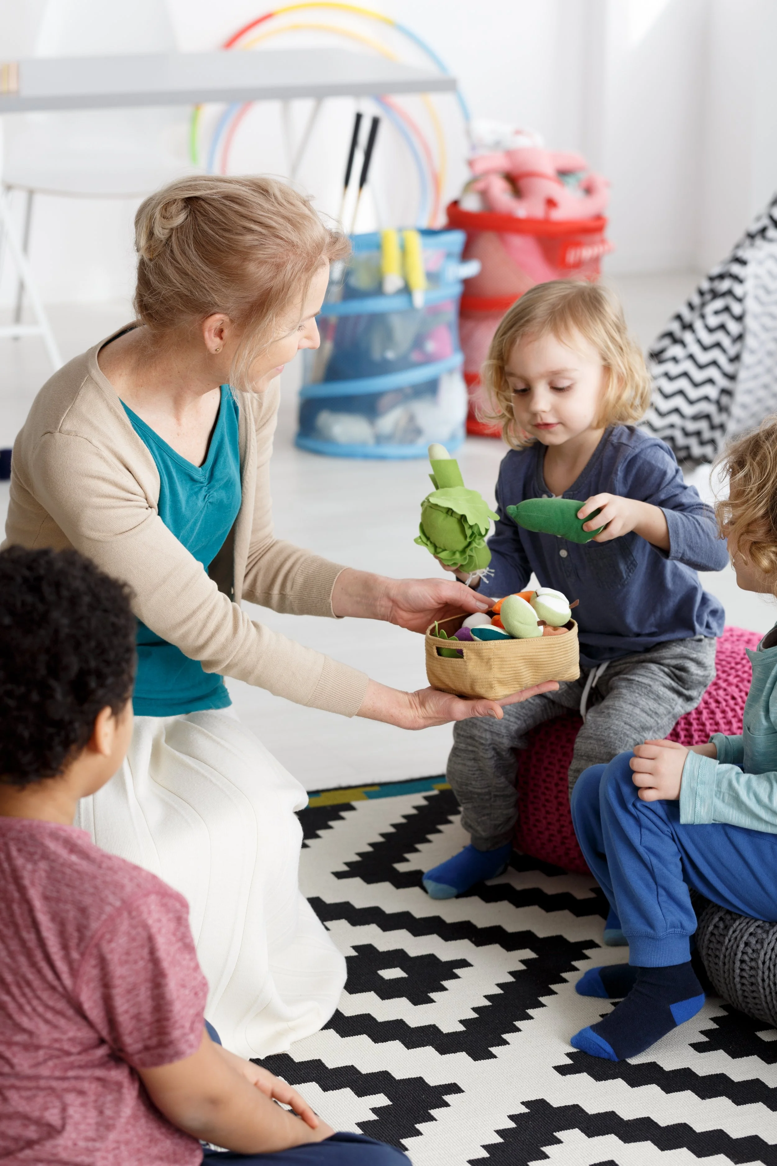 A woman is sitting on the floor with children, handing each of them a basket of toys. One girl in a blue shirt is holding a plush vegetable toy, and other children are sitting and listening on a black and white zigzag rug in a bright room with storage baskets in the background.