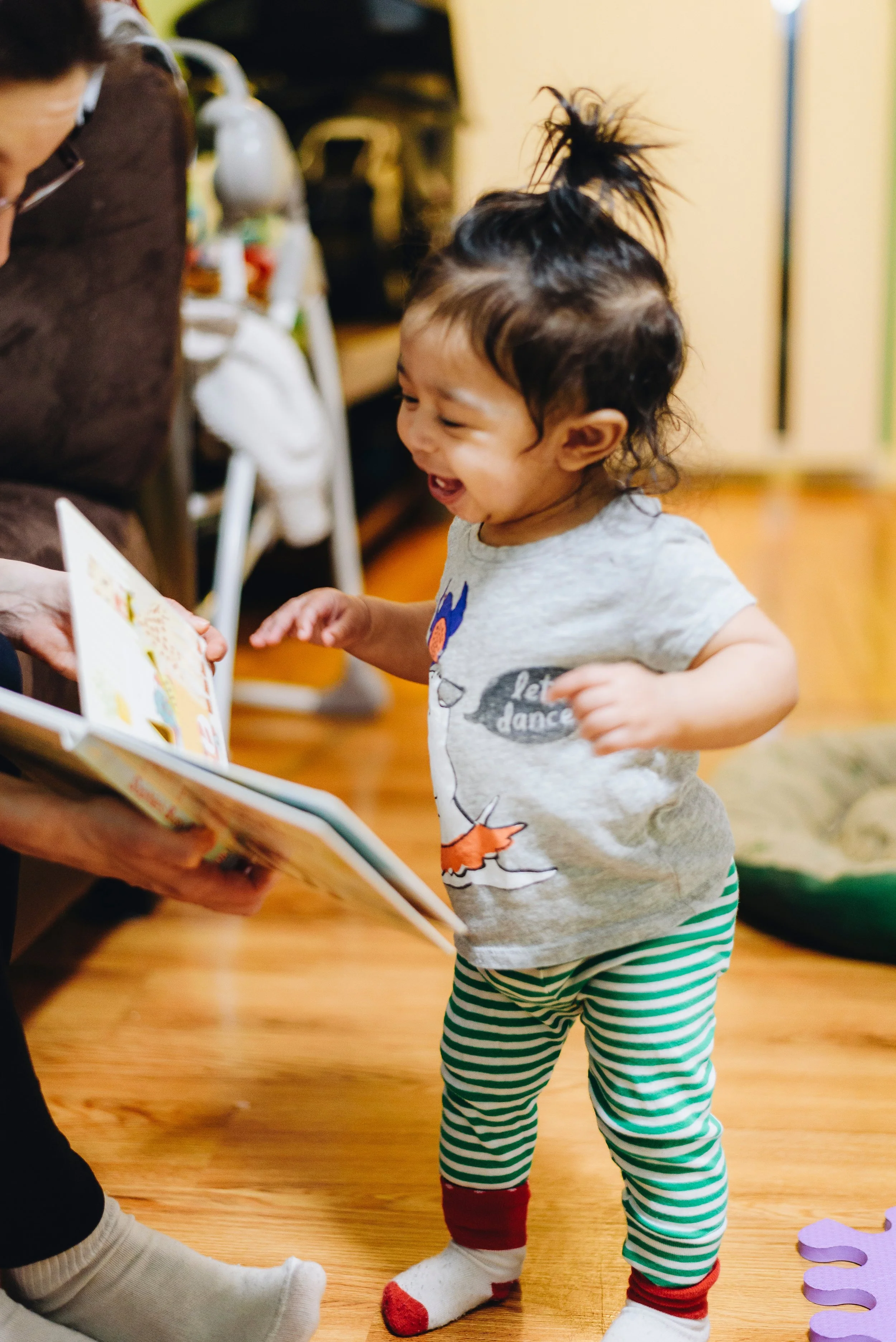 A young child with dark hair tied up in a messy bun, wearing a light gray shirt with cartoon characters and striped green and white pants, is happily looking at a book held by an adult. The child is smiling and appears to be excited, standing on a wooden floor with a purple paper chain decoration nearby.