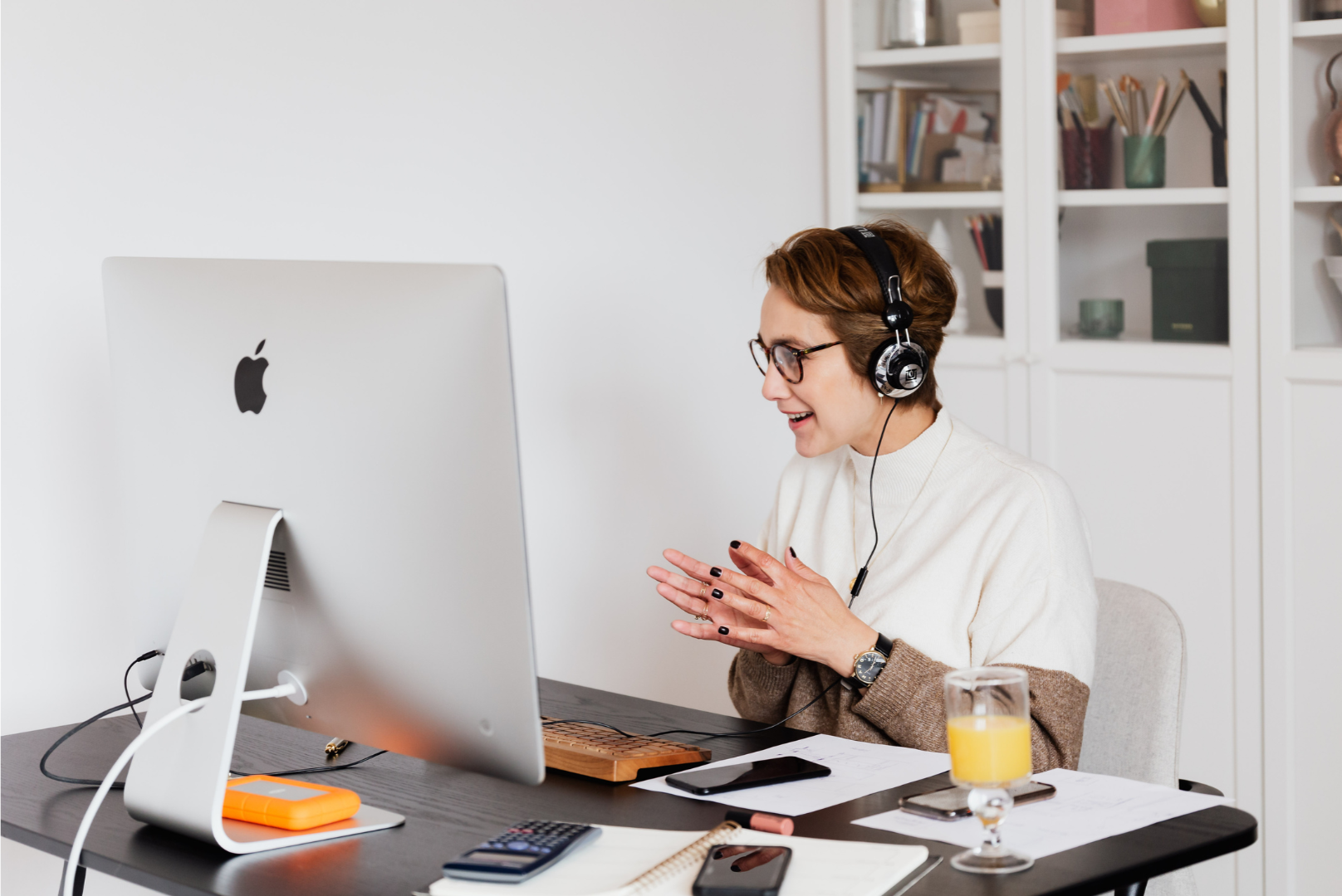 A woman sitting at a desk with a large iMac, wearing headphones, smiling with her hands clasped, in a home or office setting.