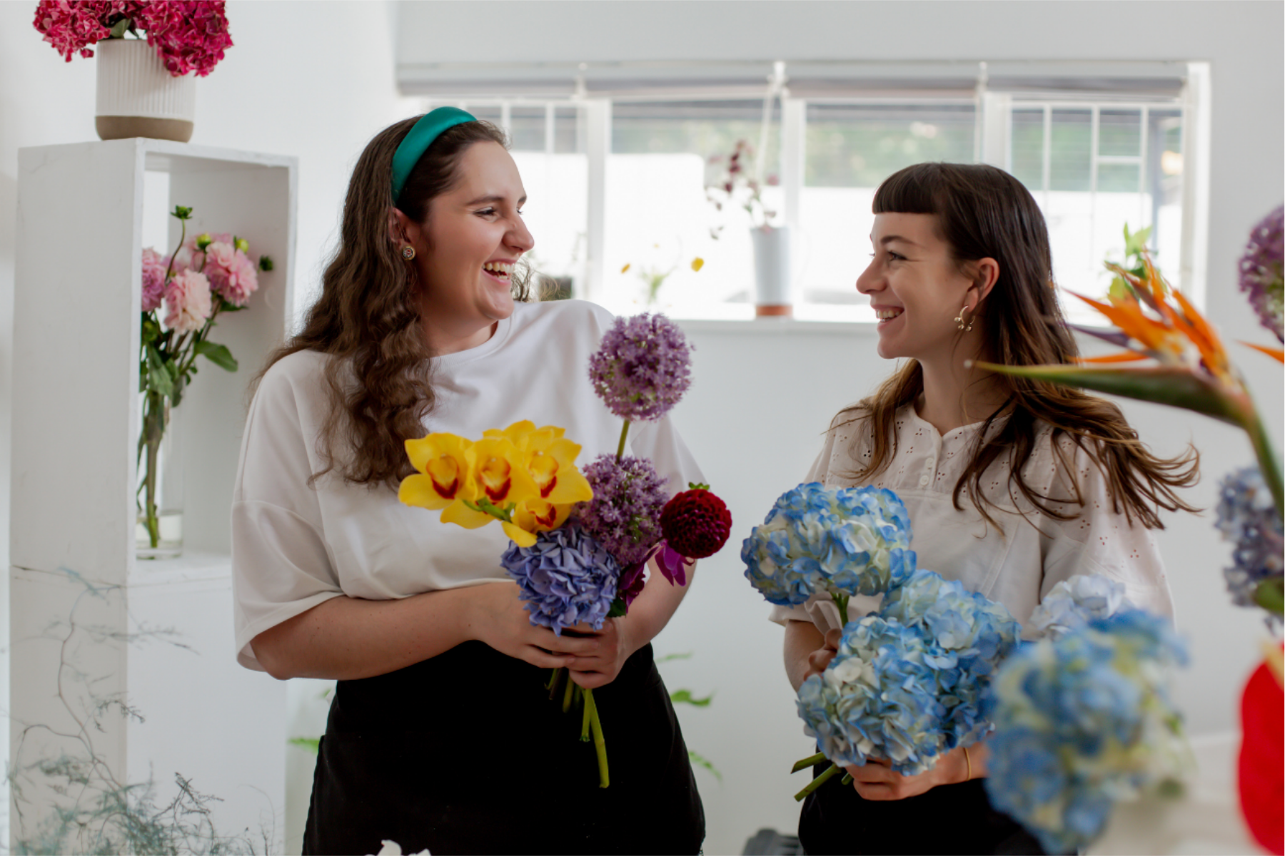 Two women smiling and holding colorful bouquets of flowers inside a bright floral shop.