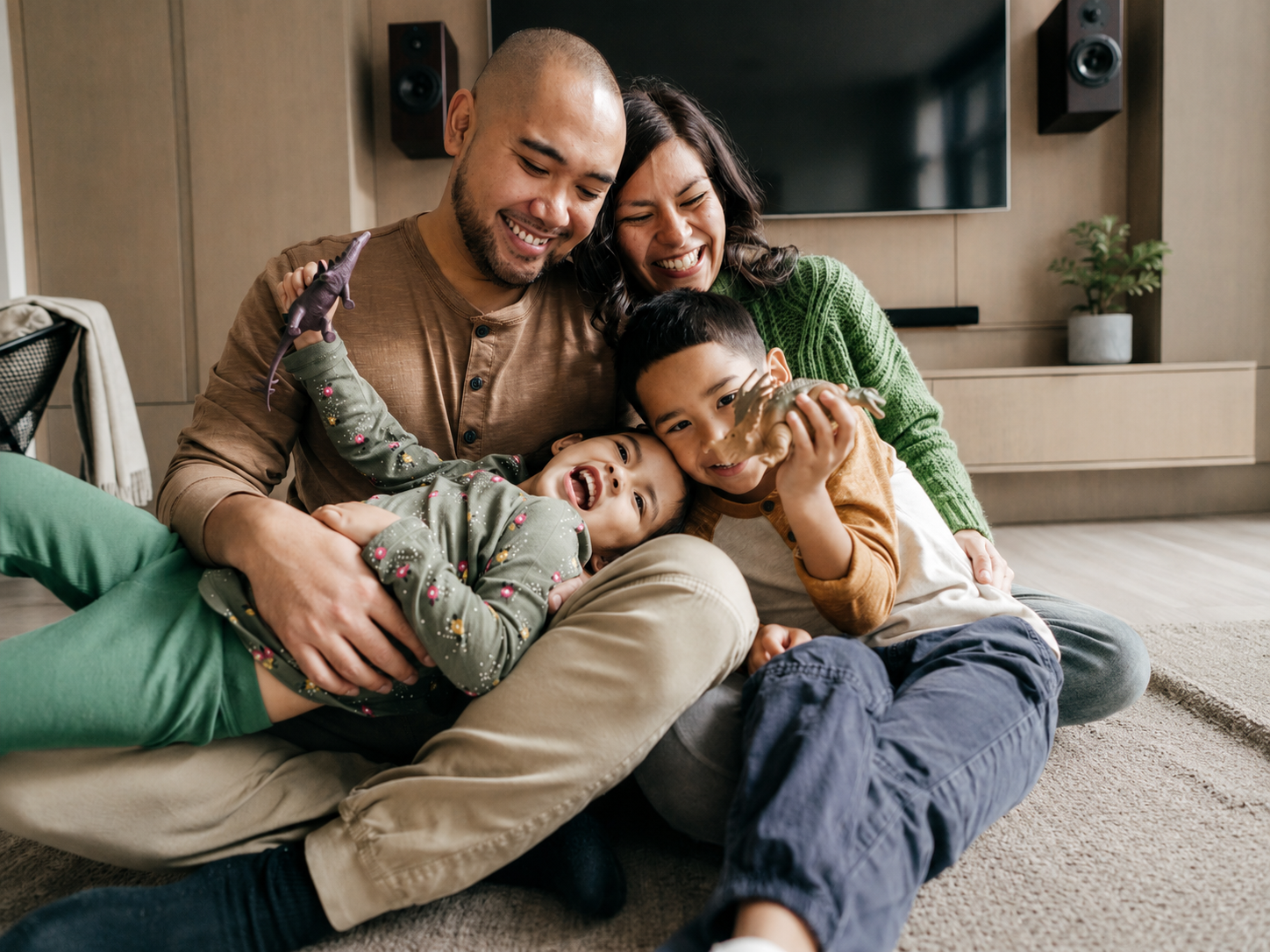 A family of four sitting on a carpeted floor in a living room, smiling and hugging each other. They are playing with plastic dinosaur toys, with a large TV and a potted plant visible in the background.