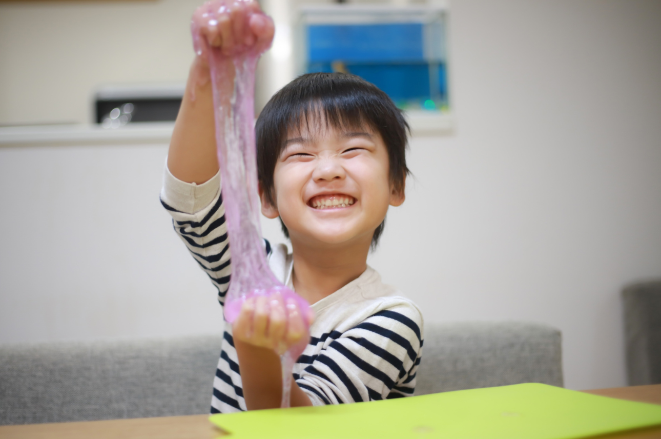 Young boy smiling and playing with pink slime at a table.