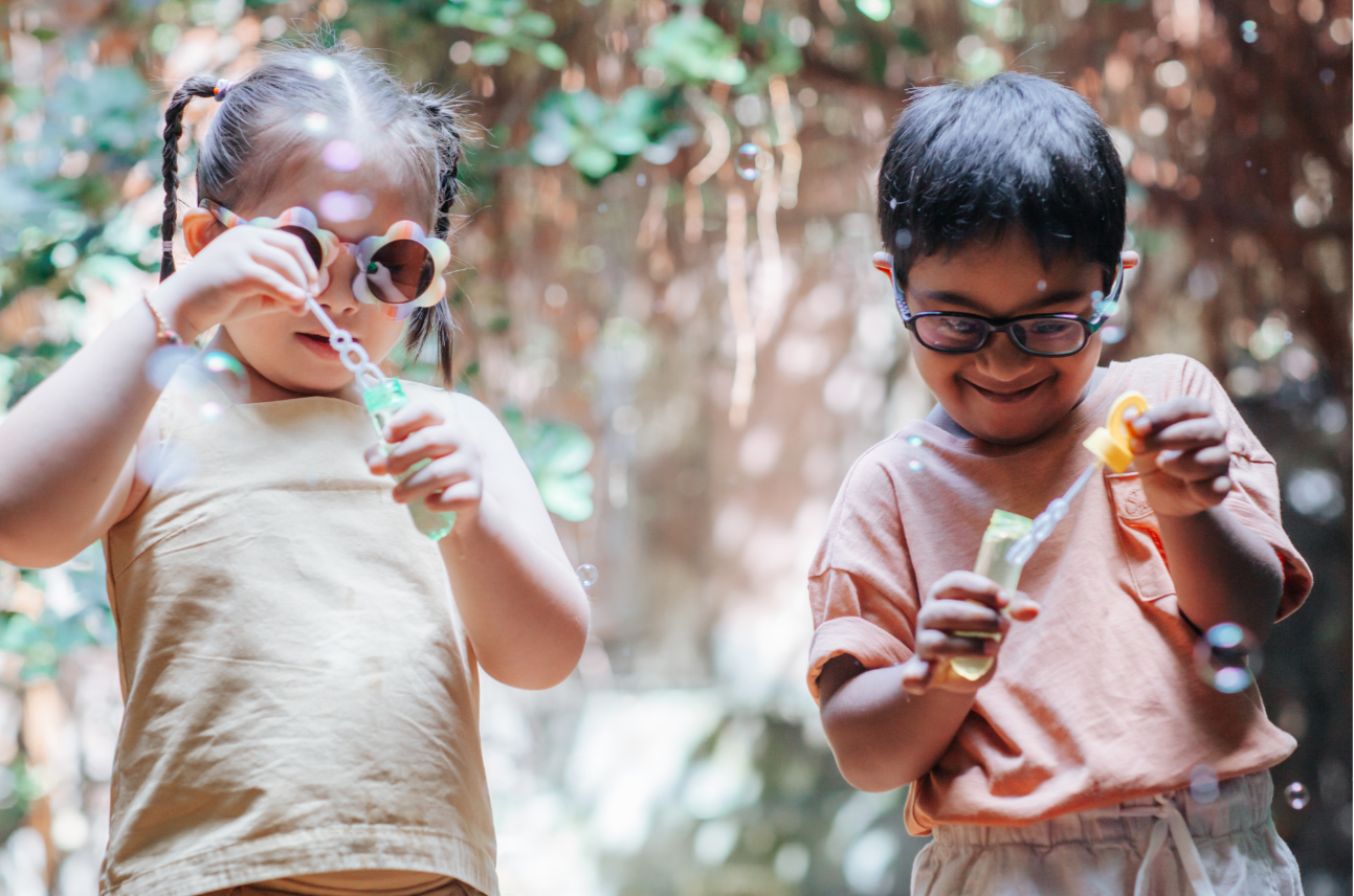 Two children outdoors, one girl with braided hair and sunglasses blowing bubbles, and a boy with glasses smiling while holding bubble solution in a park or backyard.