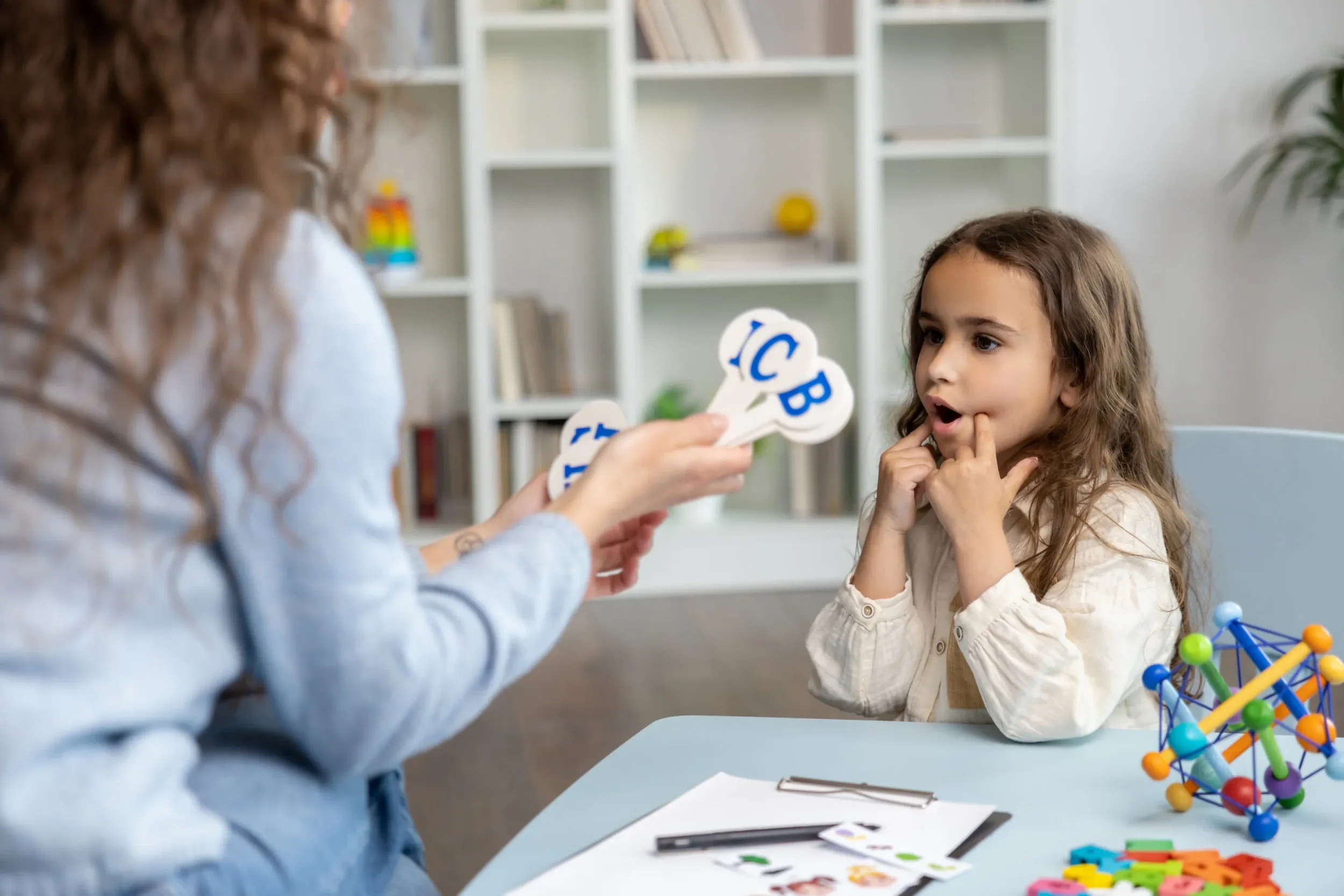 A young girl with brown curly hair sitting at a table with a teacher or adult holding up flashcards with letters to her. The girl looks focused on the flashcards, and there are colorful educational toys and materials on the table.