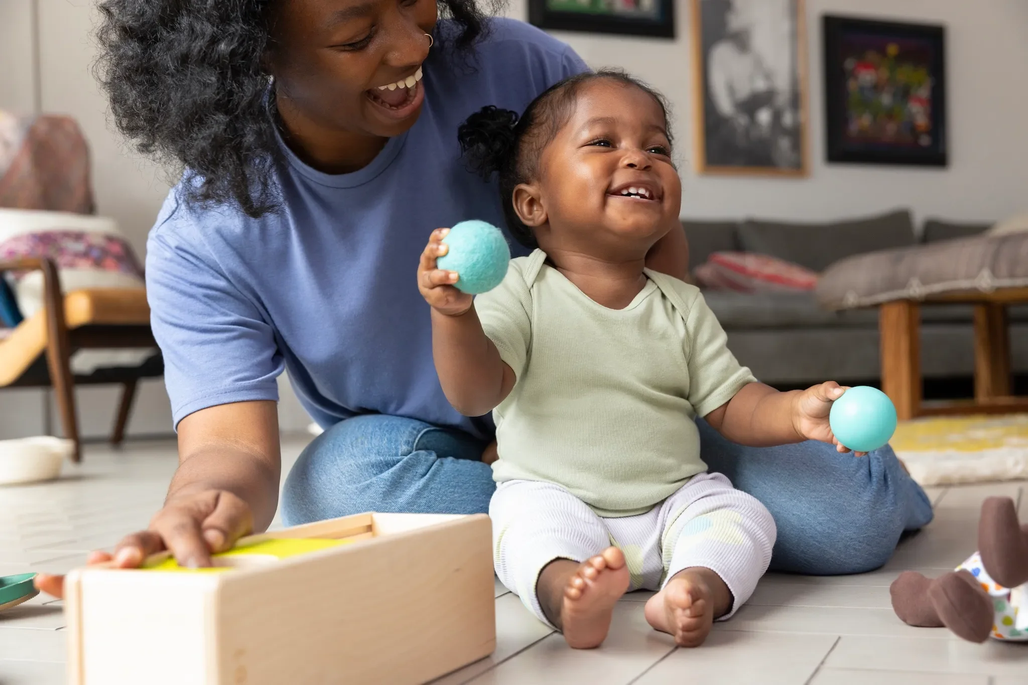 A woman and a young child playing together inside a living room. The woman is smiling and appears to be laughing, while the child is sitting on the floor holding a blue ball and smiling. The background includes furniture and framed pictures on the wall.