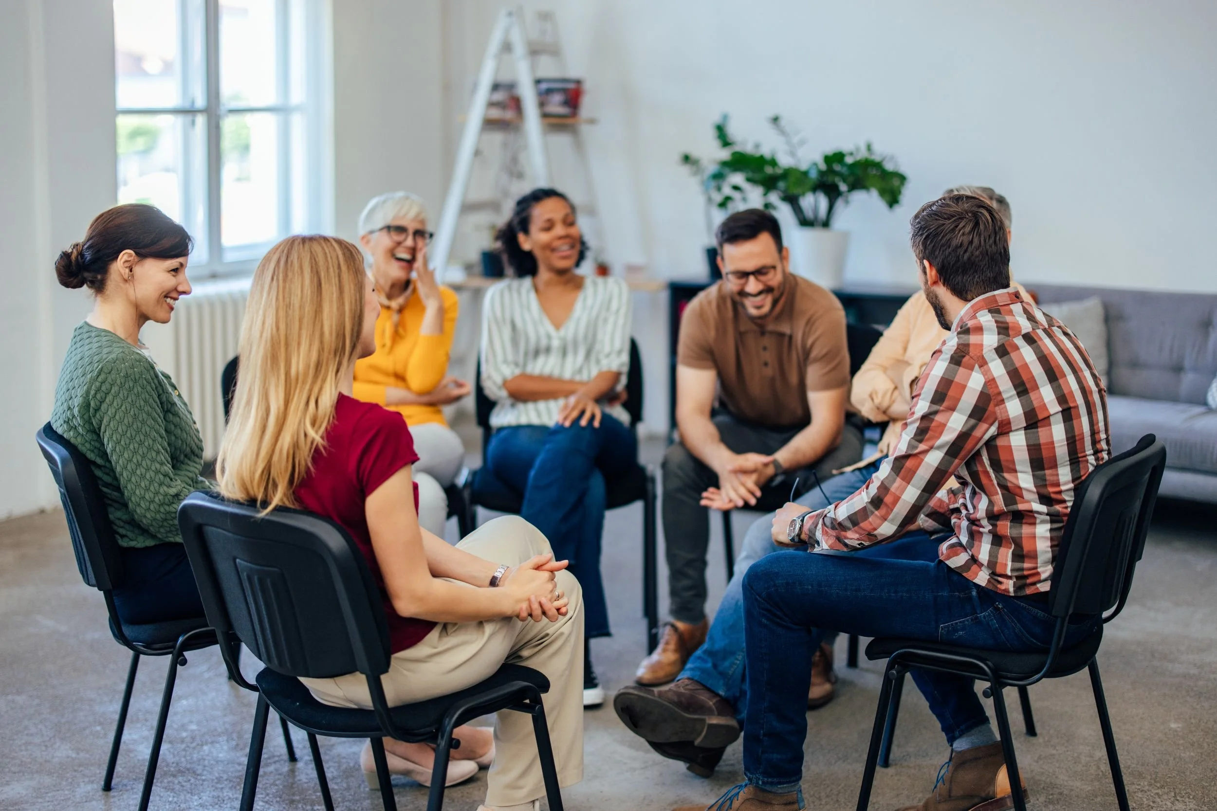 Group of seven diverse adults sitting in a circle, smiling and engaged in conversation in a bright room with large windows, plants, and casual furniture.