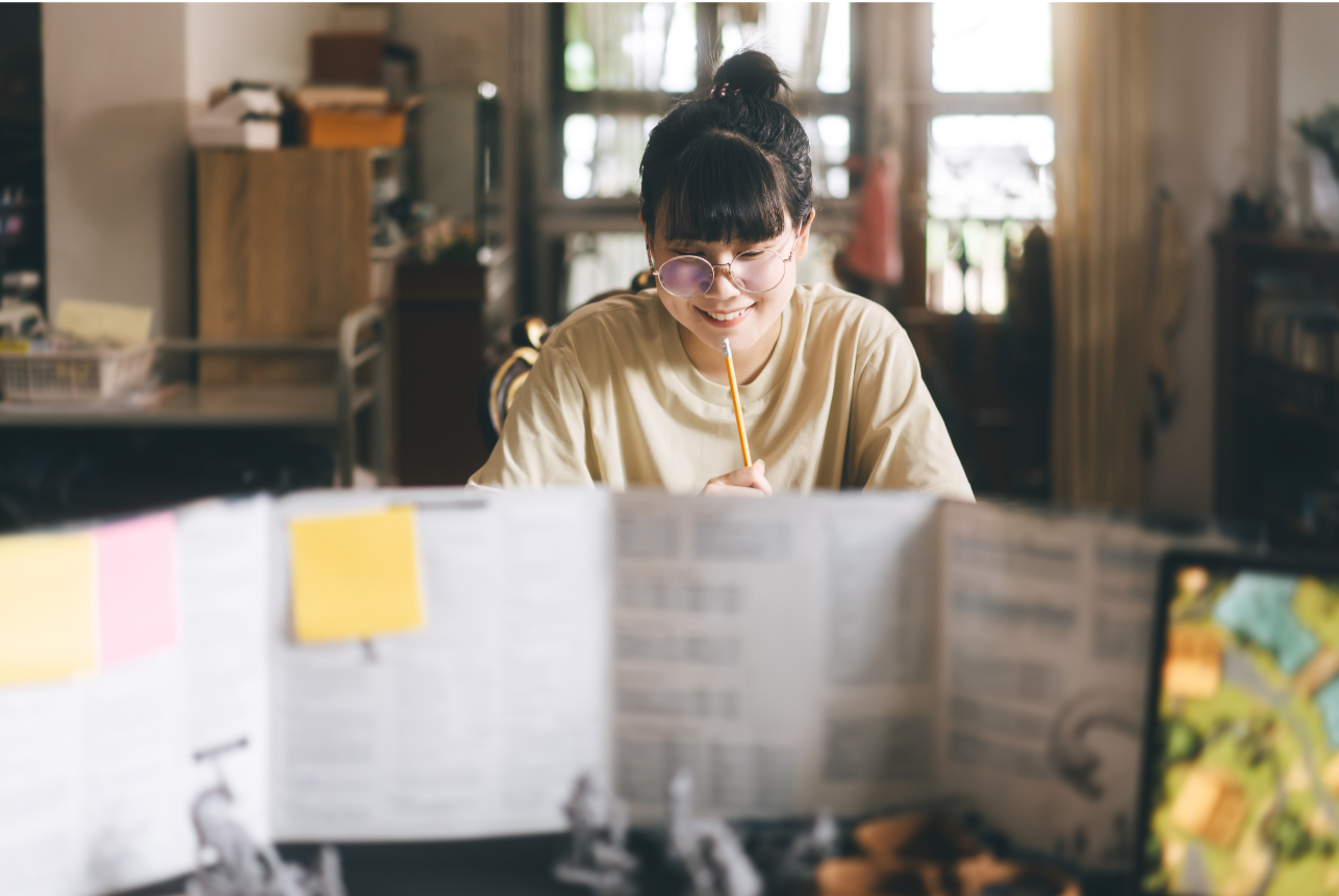 A young woman with glasses smiling while writing with a pencil at a desk, with a book open in front of her and a blurred map or game in the foreground.