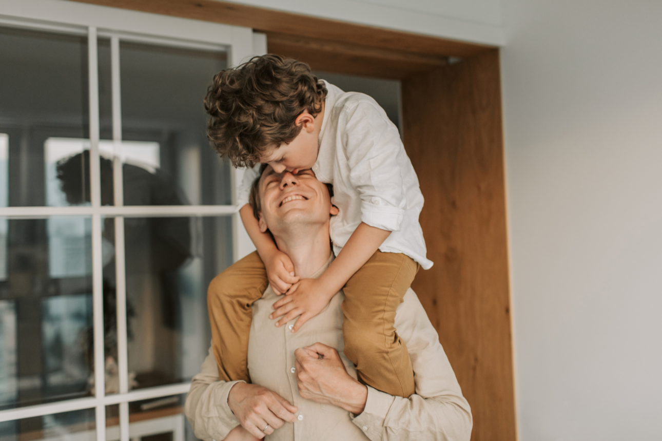 A woman lifting a young boy onto her shoulders, both smiling and sharing a joyful moment indoors.
