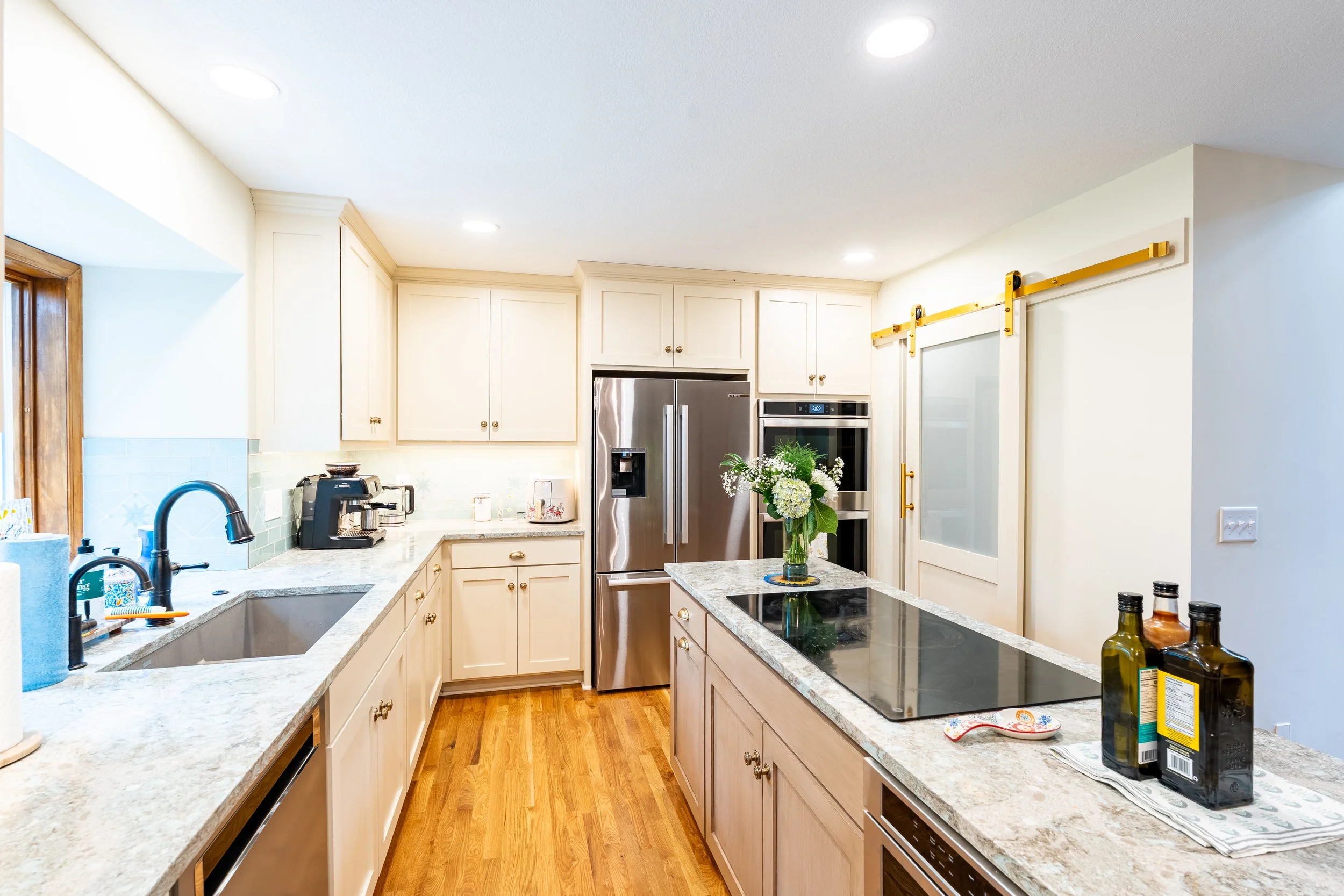 A cozy kitchen with white cabinets, a granite countertop, a stainless steel refrigerator, a vase of flowers, an induction cooktop, and various bottles and kitchen items. Recessed ceiling lights illuminate the space.