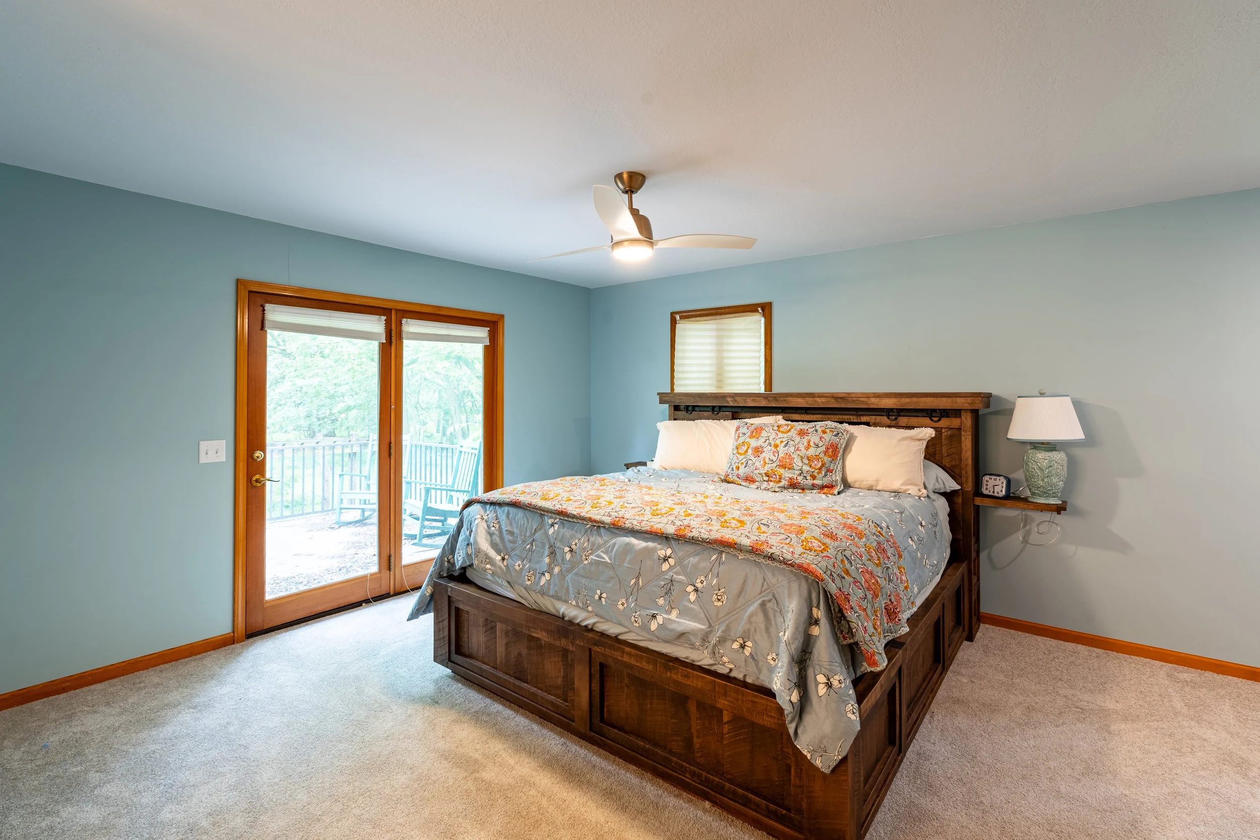 A bedroom with a wooden bed frame, floral bedding, bedside lamp, and a ceiling fan. The room has windows with blinds and a sliding glass door leading outside.