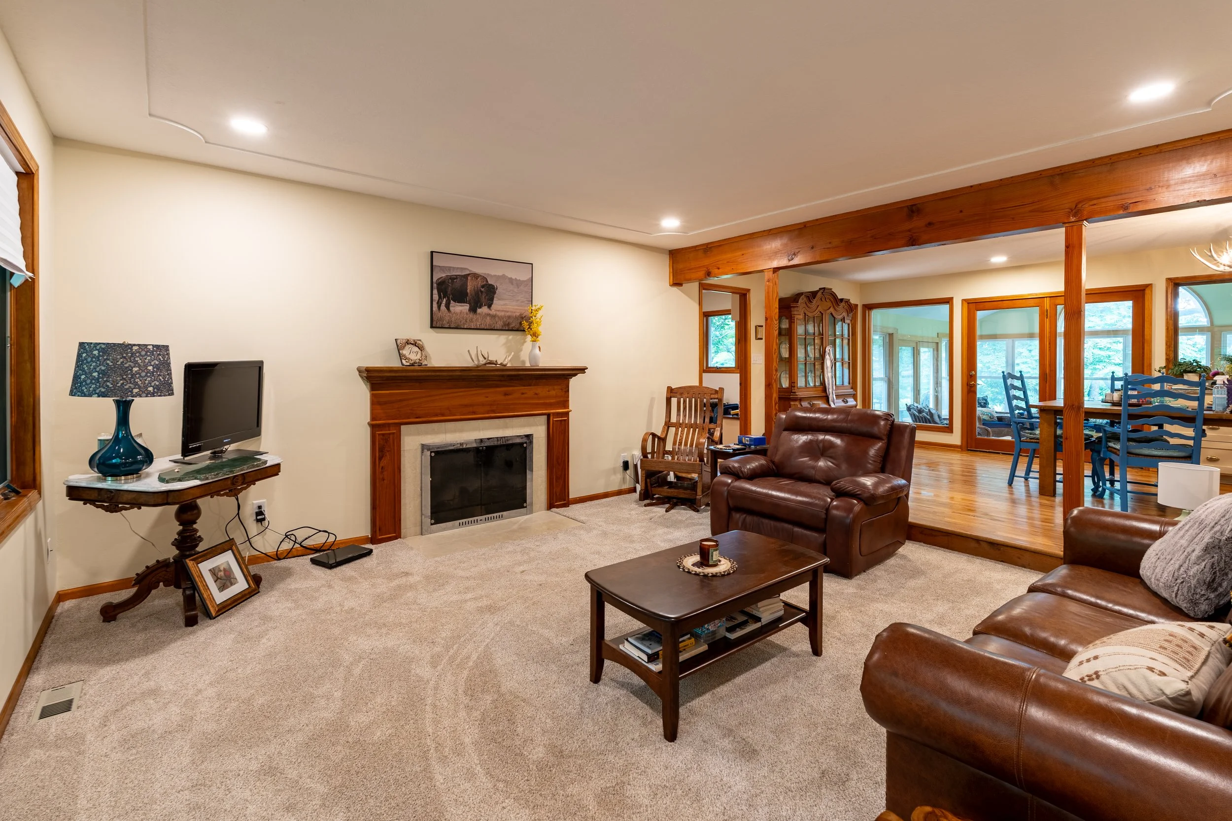 Living room with beige carpet, brown leather sofas, wooden furniture, a fireplace, and a wall art of bison, with windows showing greenery outside.