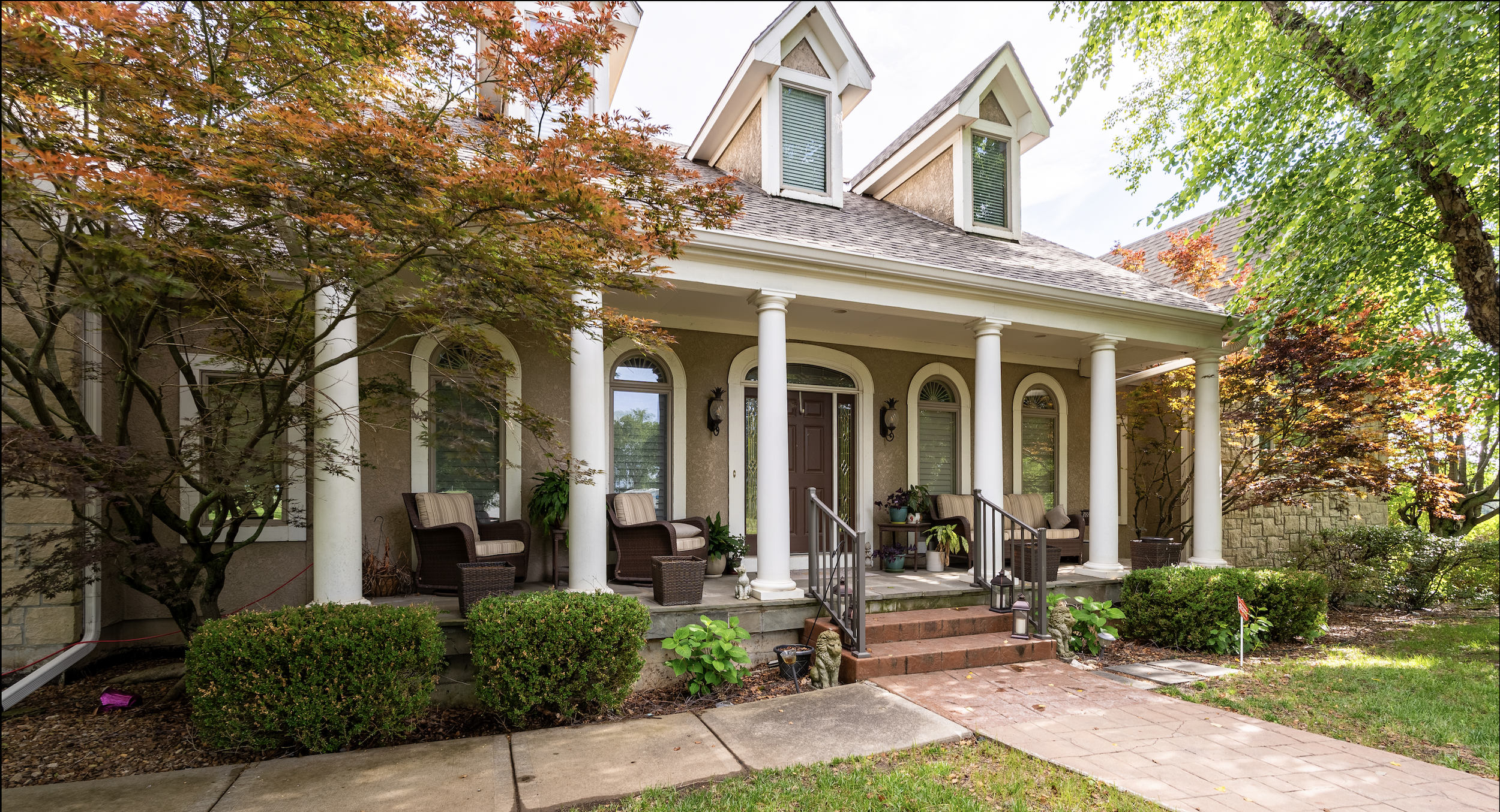 Front porch of a house with columns, chairs, plants, and trees with autumn-colored leaves.