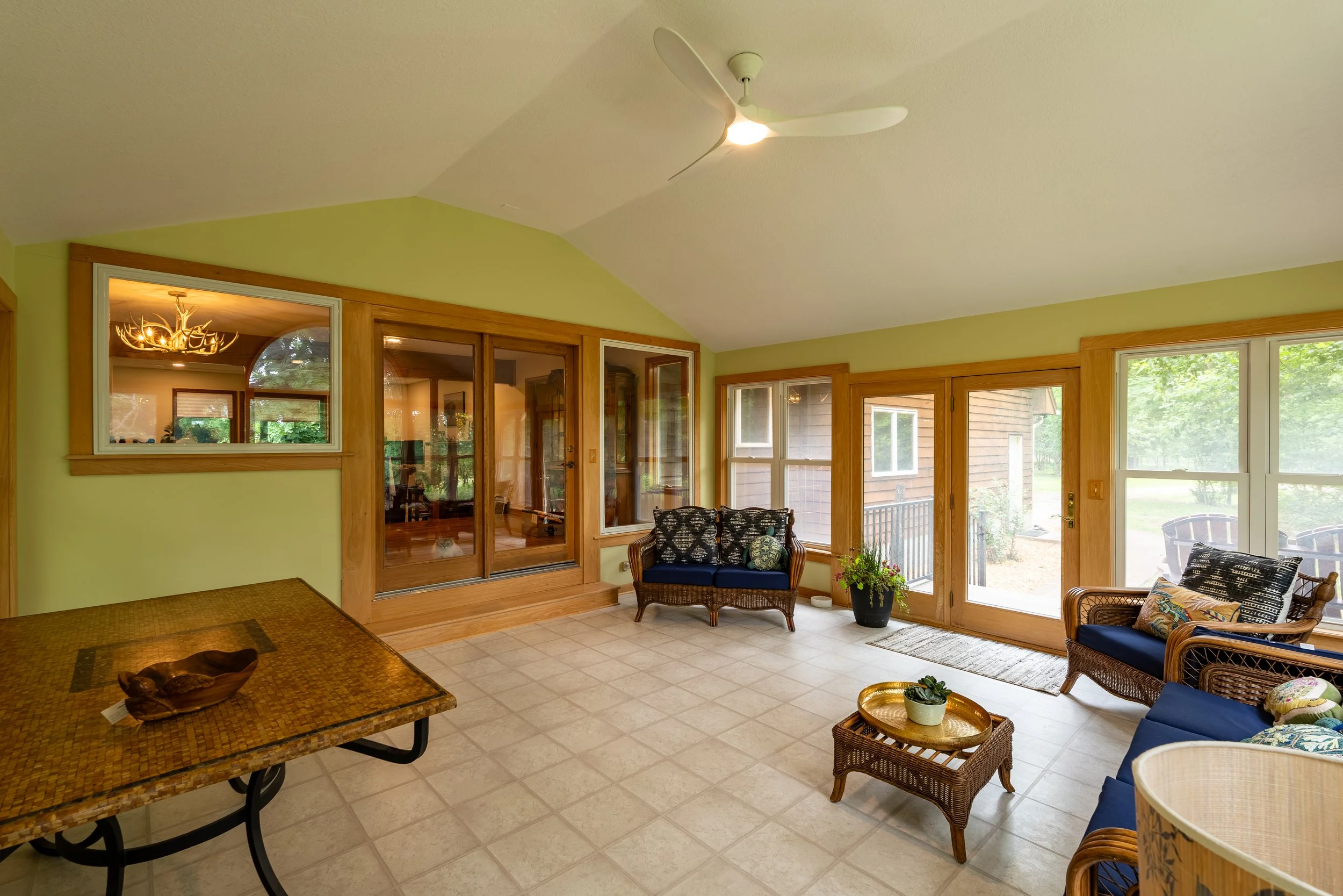 A bright sunroom with large windows and a ceiling fan. There are wicker chairs with blue cushions, a small table with a plant, and a sliding glass door leading outside. The room has light-colored tiled flooring and green walls.