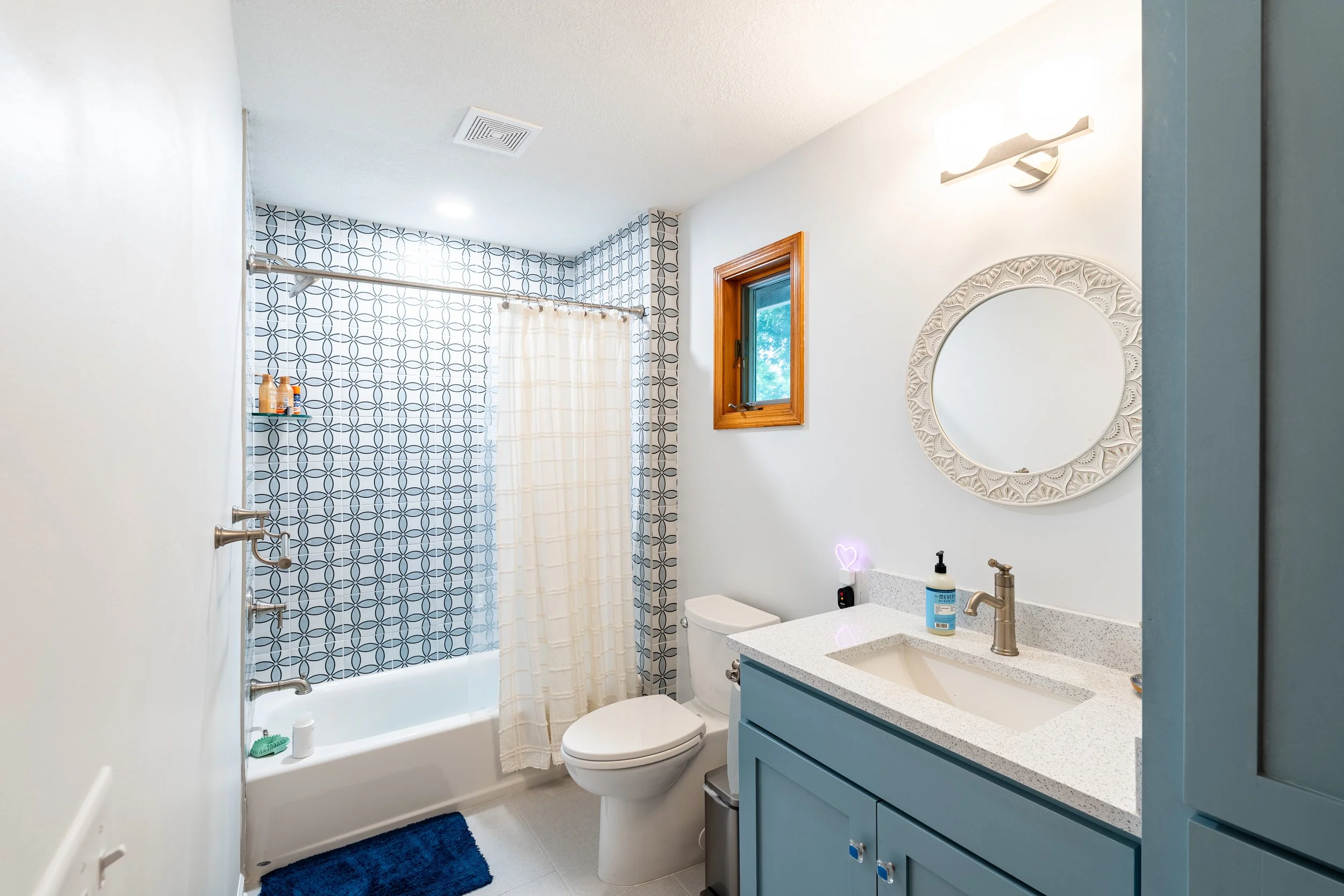 Bathroom with white walls, a bathtub with a shower curtain, a toilet, a vanity with a blue cabinet, a sink, a mirror, a window, and a small blue rug.