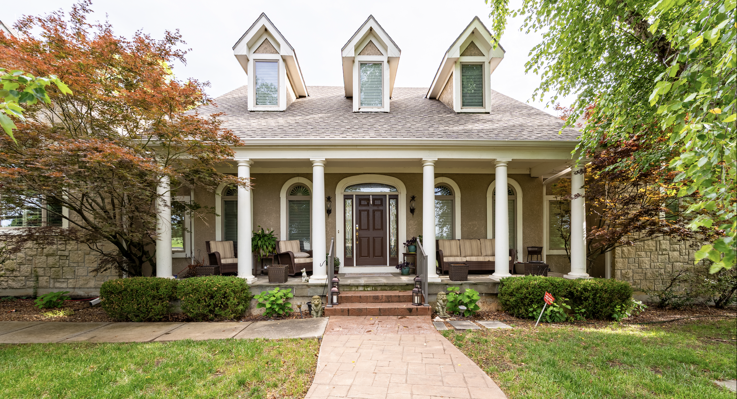 Front view of a house with a porch, potted plants, benches, and a brick pathway leading to the entrance, surrounded by trees and shrubs.