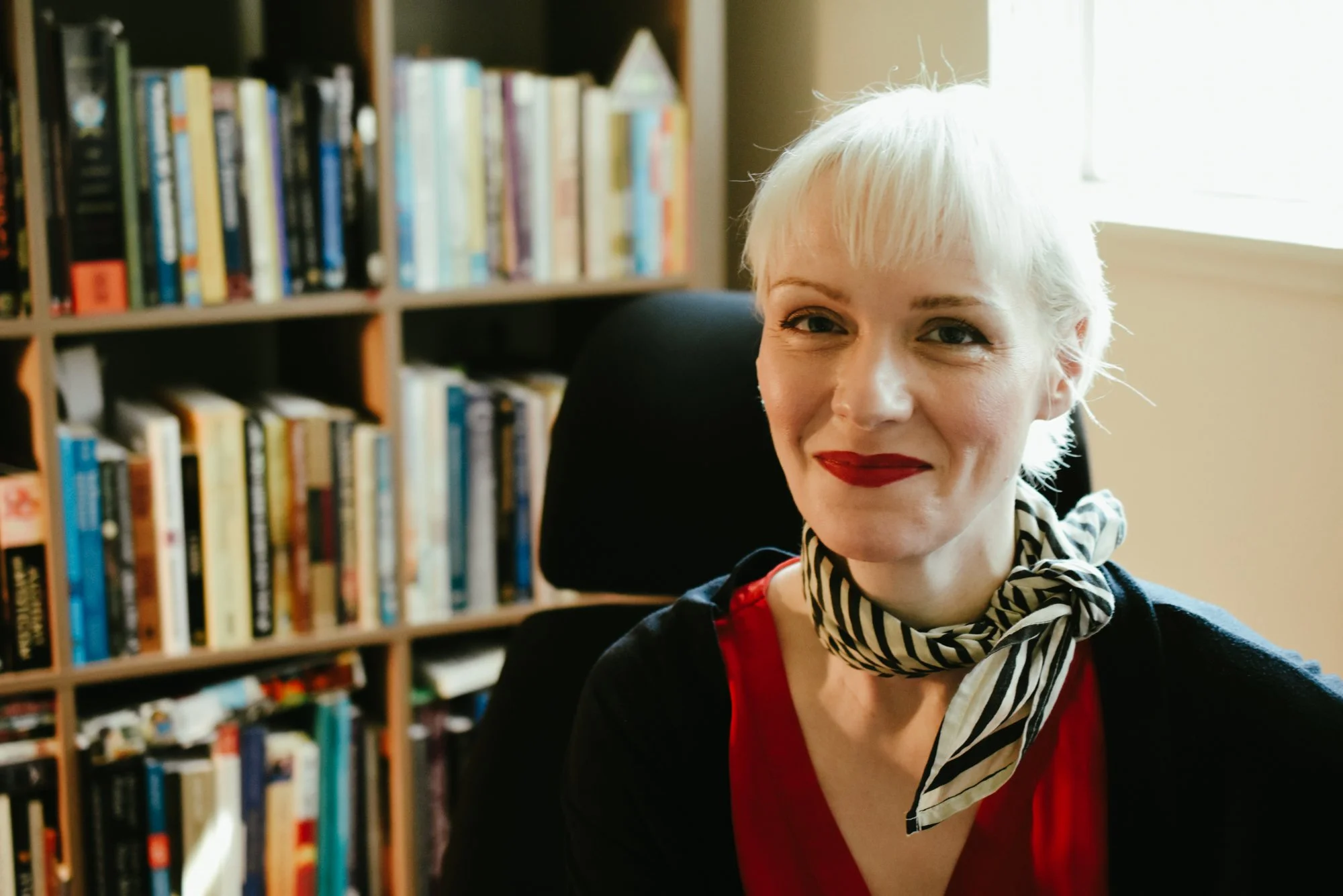 Madeline, with short  blonde hair and red lipstick smiling at the camera, sitting in front of a bookshelf filled with books.