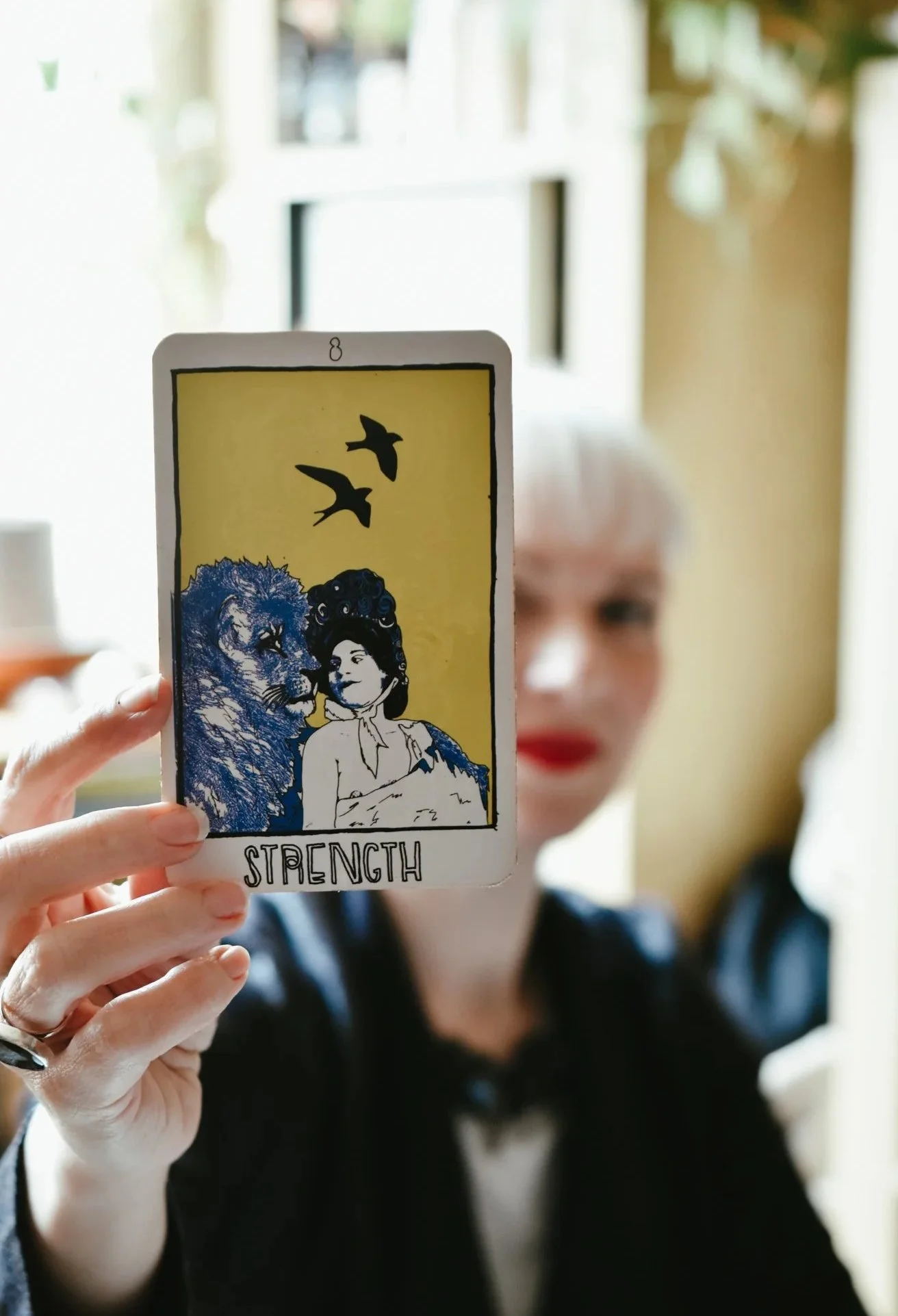 Madeline blurred in the background, holding a tarot card of Strength with an illustration of a woman and a lion, two birds flying above.