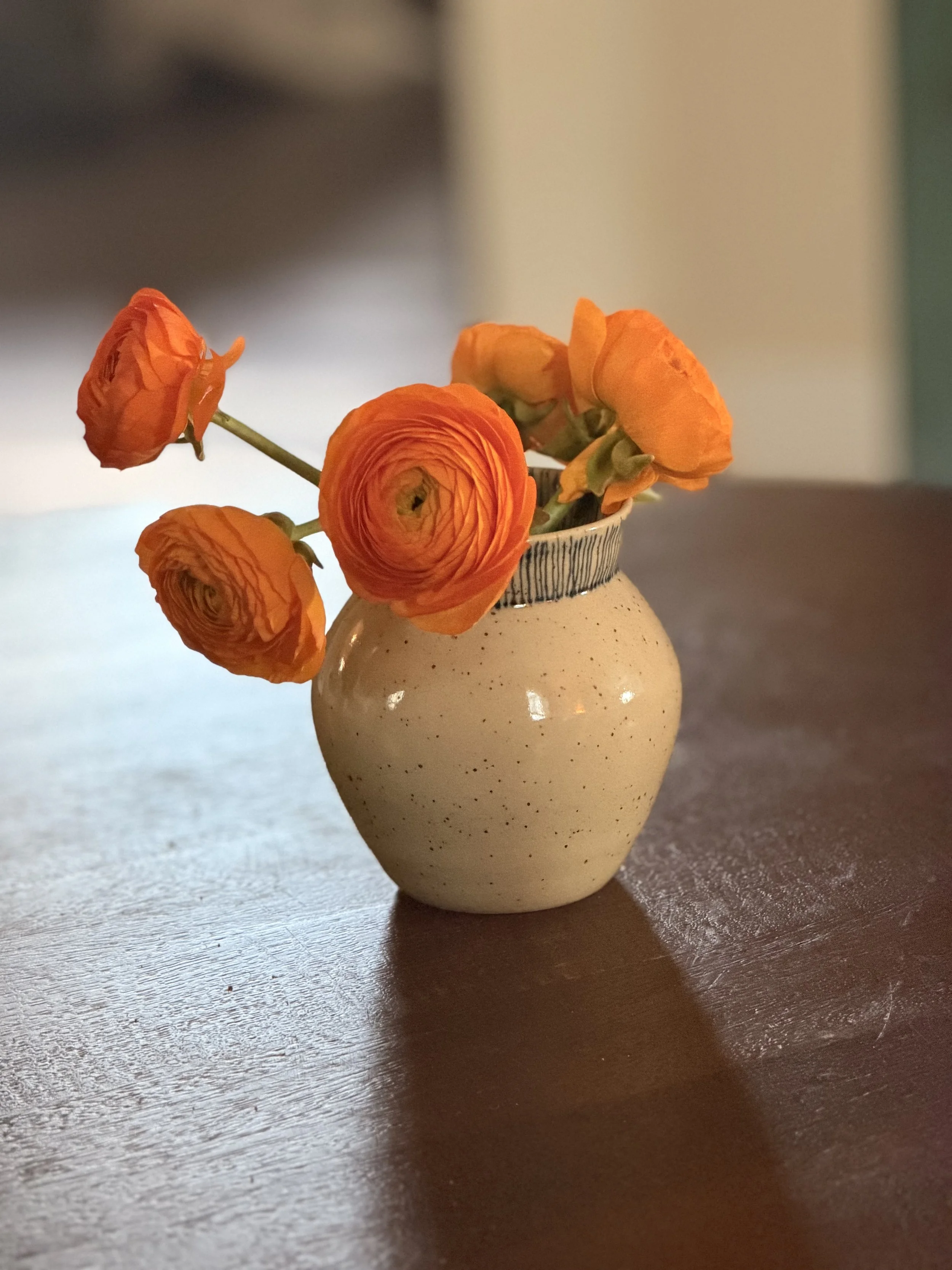 A small beige ceramic vase with a narrow opening holding orange ranunculus flowers on a wooden surface.