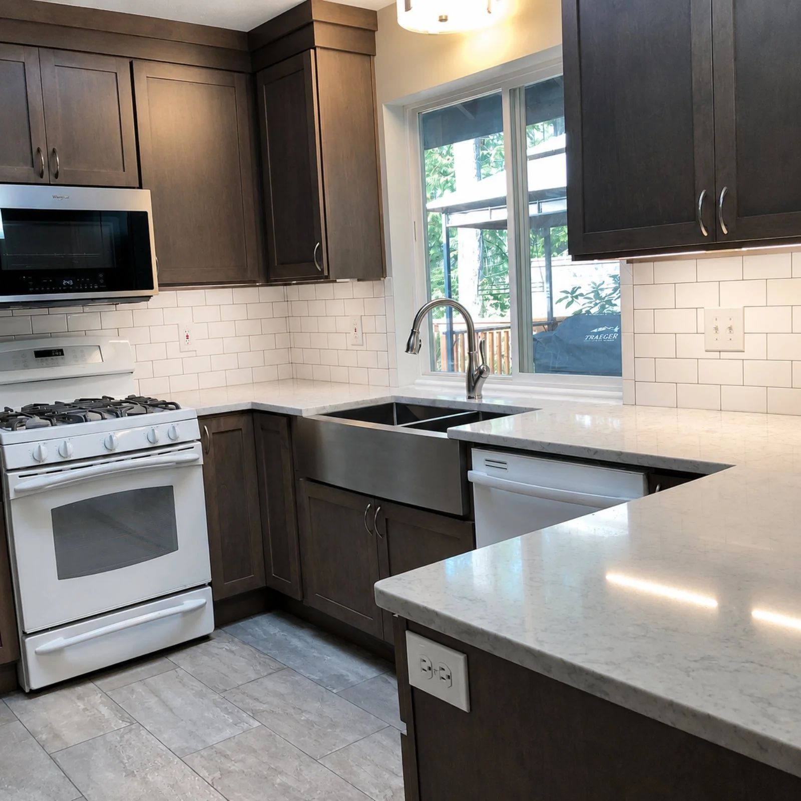 Modern kitchen with dark wood cabinets, white subway tile backsplash, white stove, microwave, and a stainless steel sink by a window overlooking a porch and trees.