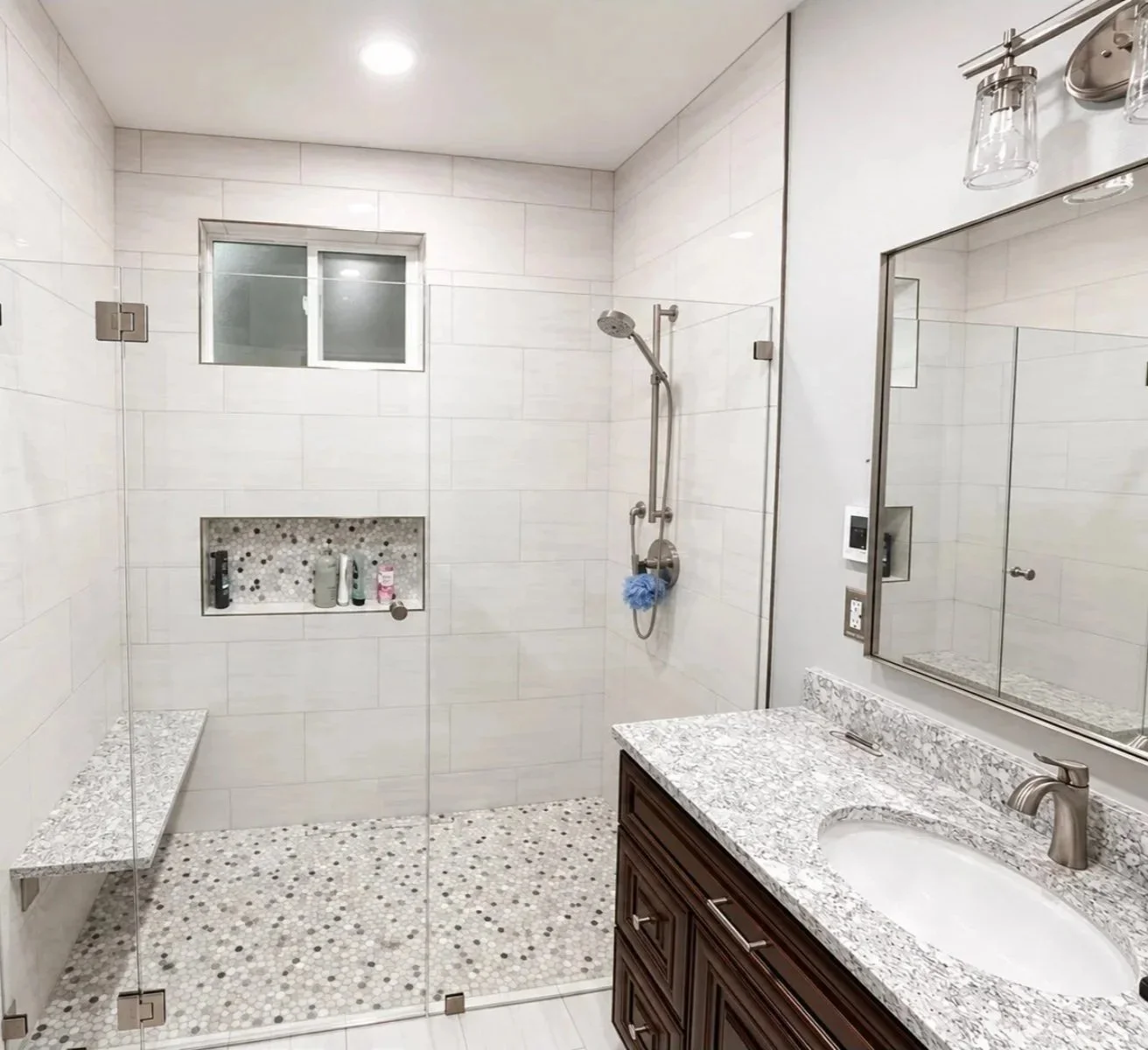 Modern bathroom with a glass-enclosed shower, wall-mounted showerhead, built-in shower niche, granite vanity countertop with a sink, mirror, and dark wooden cabinetry.