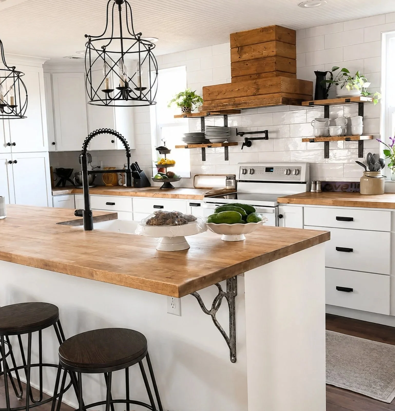 A modern kitchen with white cabinets, wooden countertops, black fixtures, and black stools around a wooden island with two bowls of vegetables. Open shelves hold plates, cups, and plants, with a range hood and white tiled backsplash behind the stove.