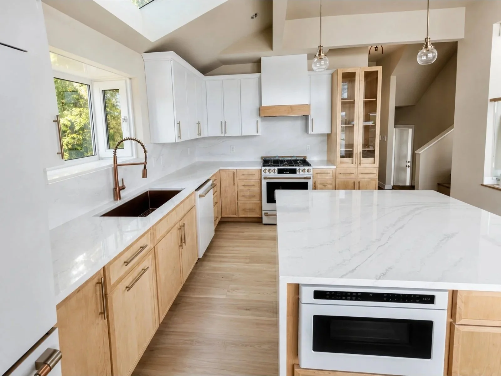 Modern kitchen with white and light wood cabinets, a white marble countertop, a copper faucet, a window with greenery outside, a white stove and oven, and pendant lights hanging from the ceiling.